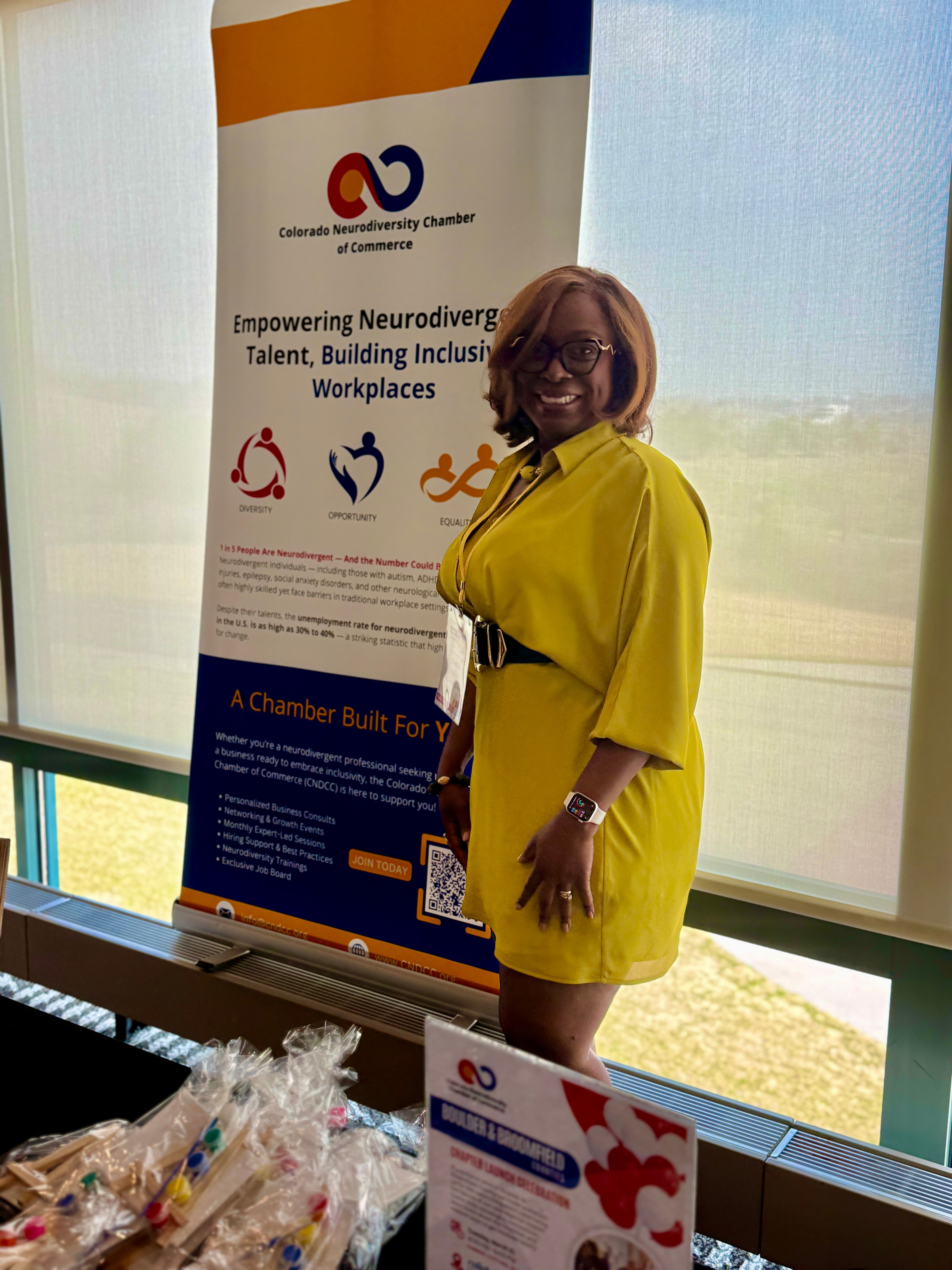 Woman in yellow dress standing next to a banner, smiling, with a table of items in front, large window behind, bright daylight.