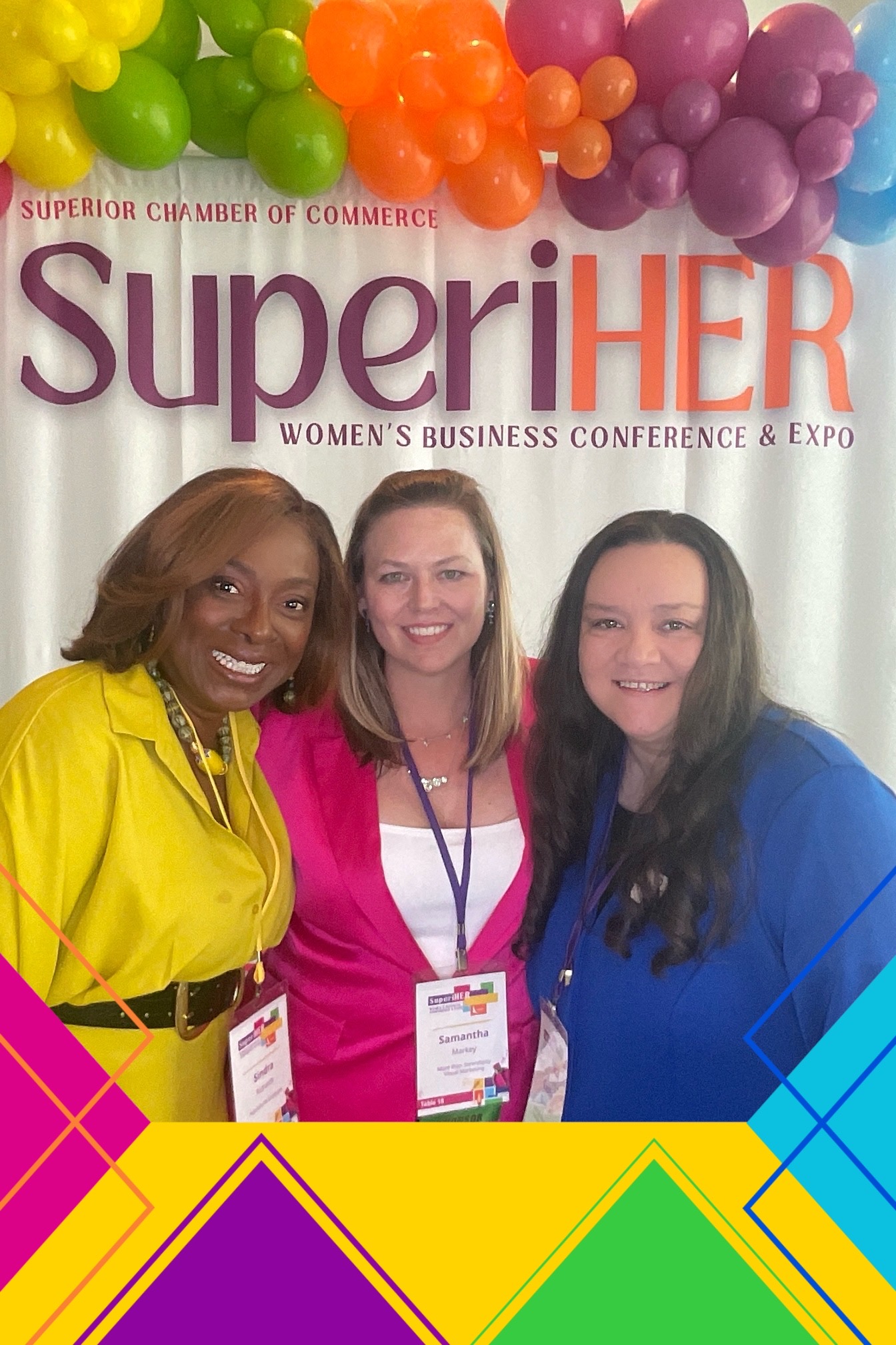 Three women smiling at a conference, wearing colorful clothing and name tags, with balloons in the background.
