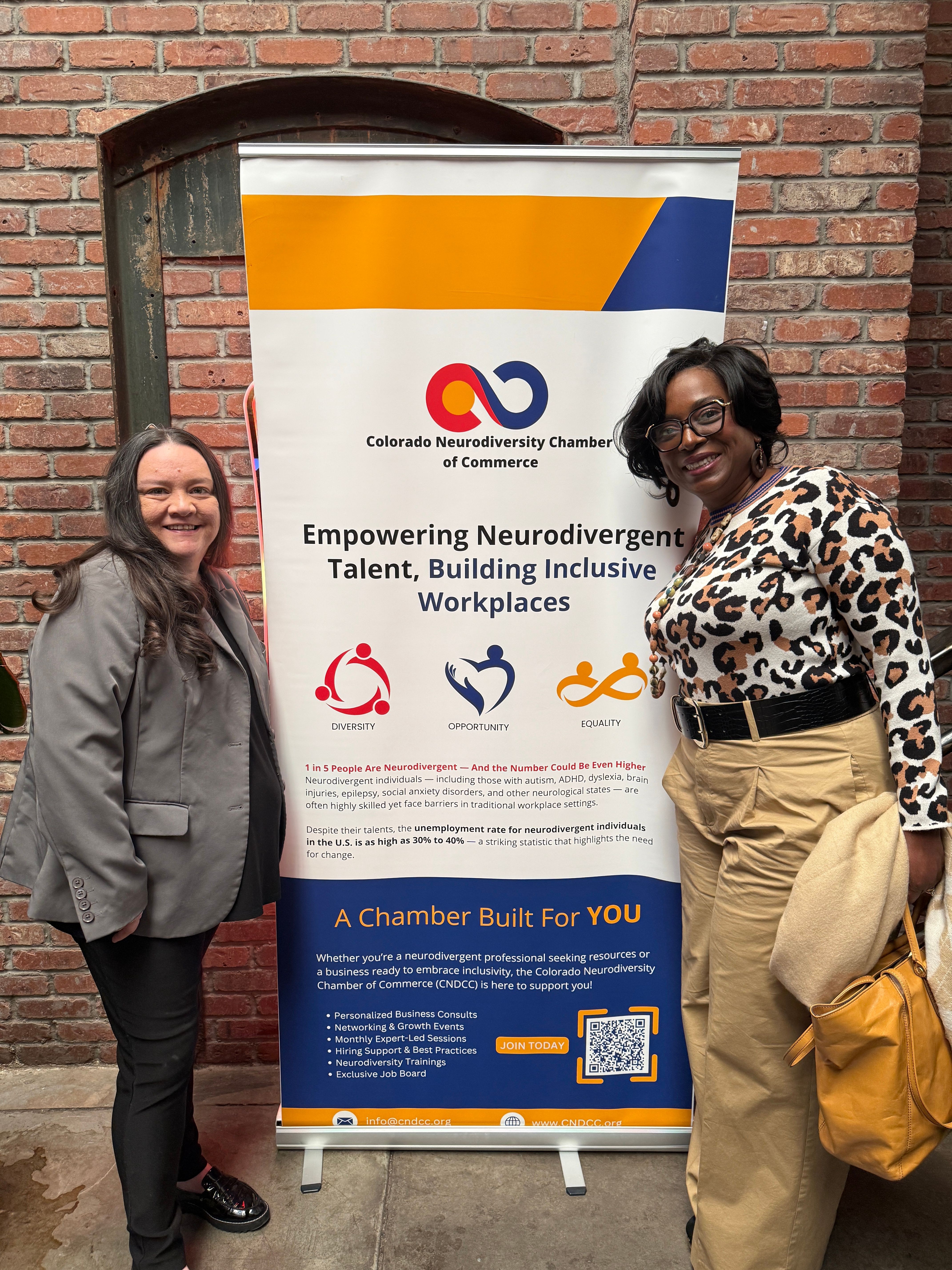 Two women standing next to a vertical informational banner against a brick wall, one wearing a leopard print top and beige pants, the other in a gray jacket and black pants.