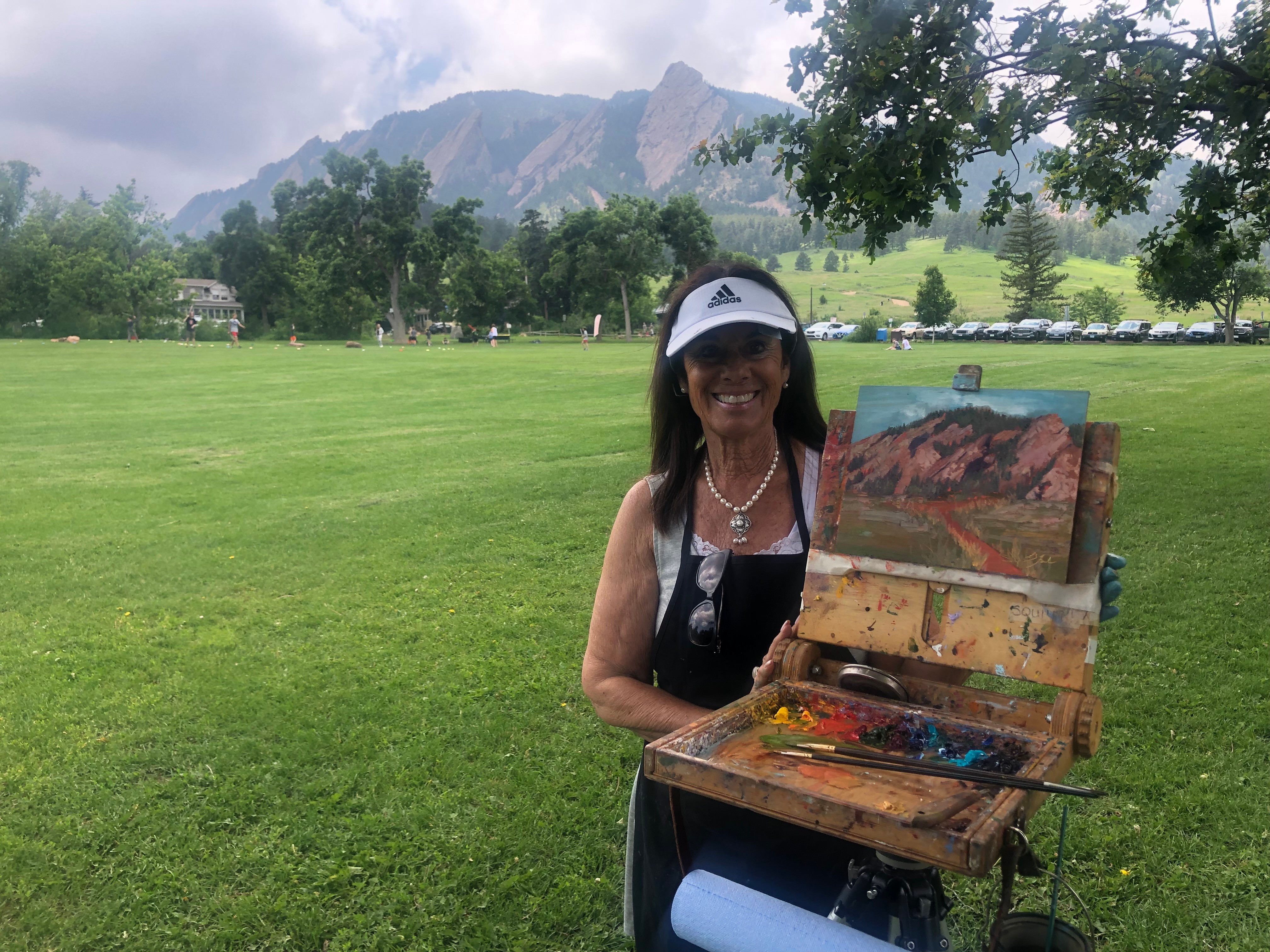 Woman outdoors holding an open painting box with landscape artwork, trees, mountains, and cloudy sky in background.