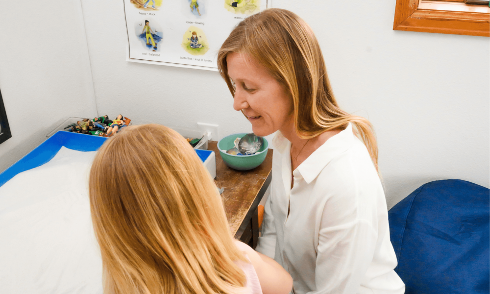A woman and a young girl sit at a desk in a room with a poster about body parts on the wall, a window, and a blue bean bag. The woman is smiling at the girl, who is looking at a book.
