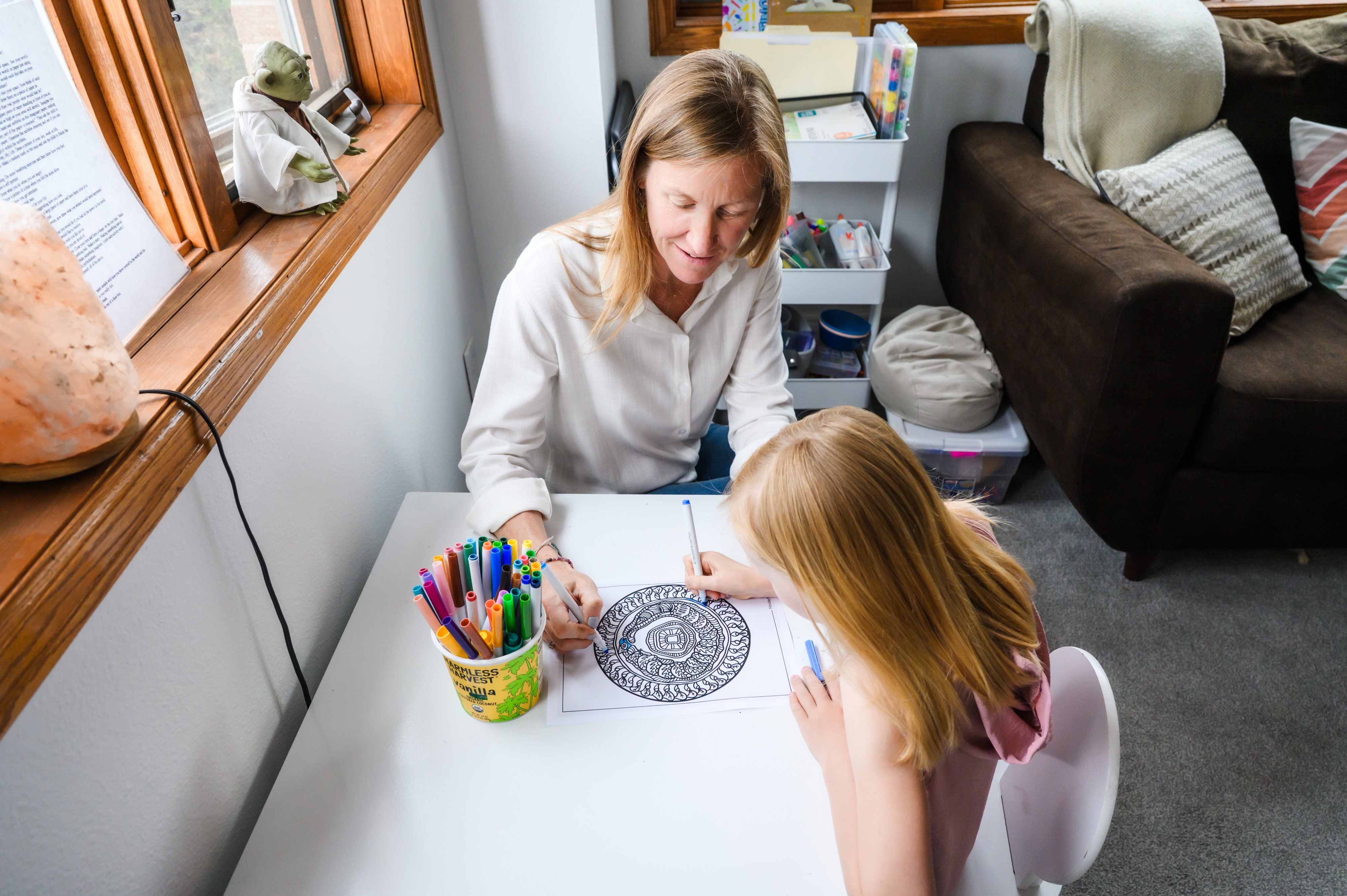 Woman and girl sitting at a white table with colorful pens, engaging in a drawing activity, in a cozy room.