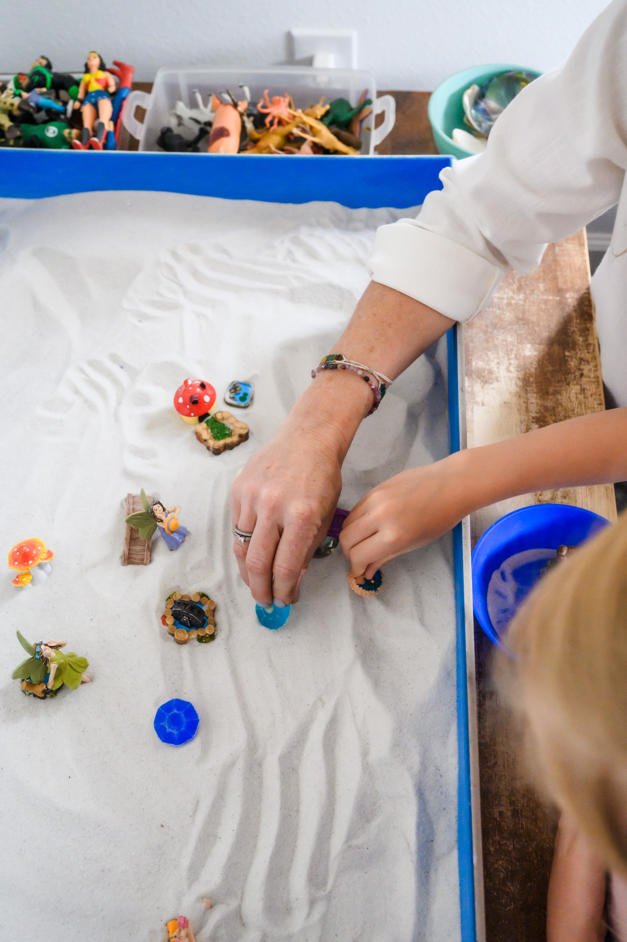 Person's hands playing with small toys on a white surface, with a child nearby and toy storage in background.