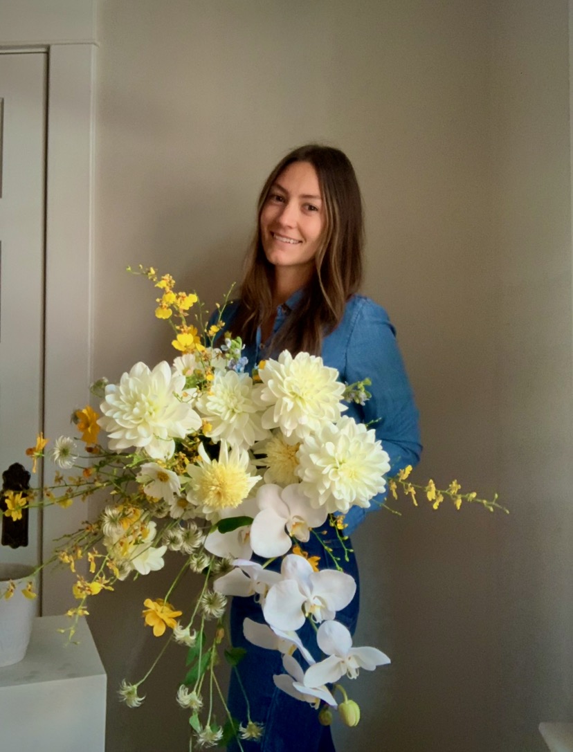 Young woman with long brown hair smiling, holding a large bouquet of white, yellow, and purple flowers, standing indoors.