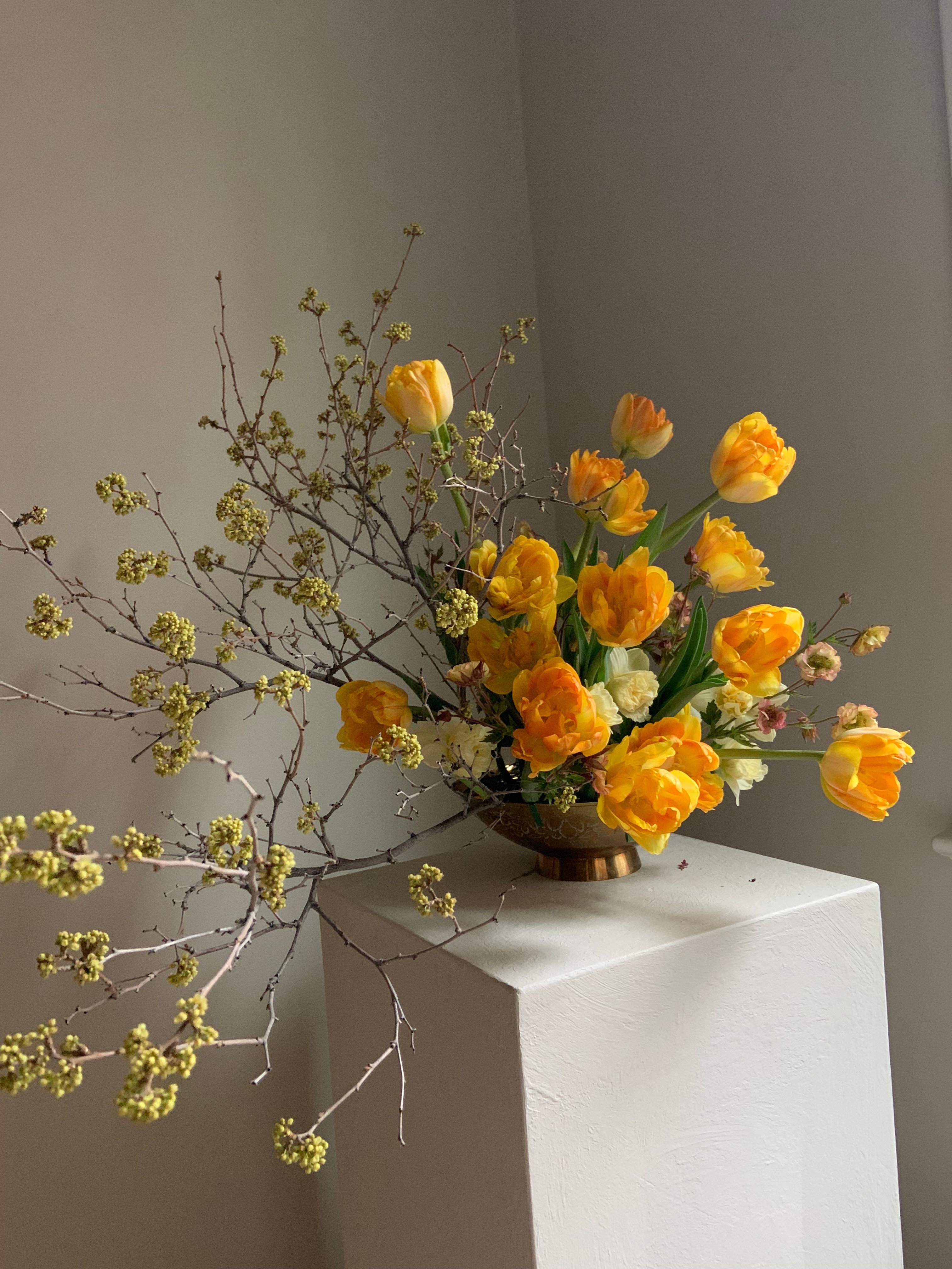 Yellow tulips and branches with small yellow flowers in a brown vase on a white pedestal, against a plain wall.