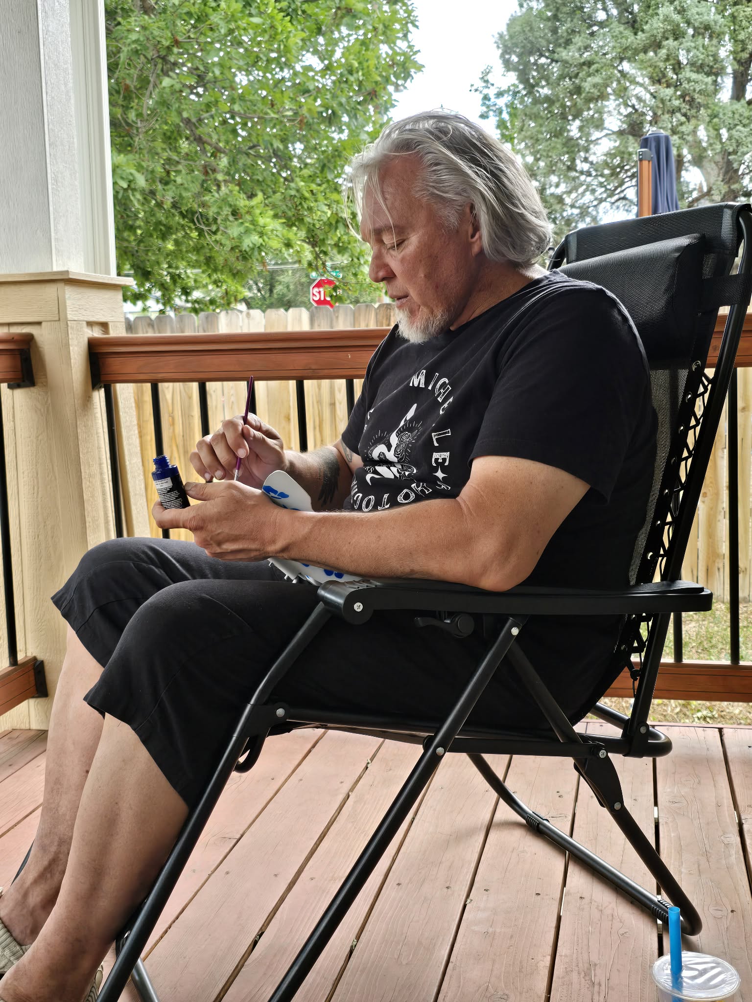 Older man with gray hair sitting on a porch, holding a small object, with trees and sky in the background.