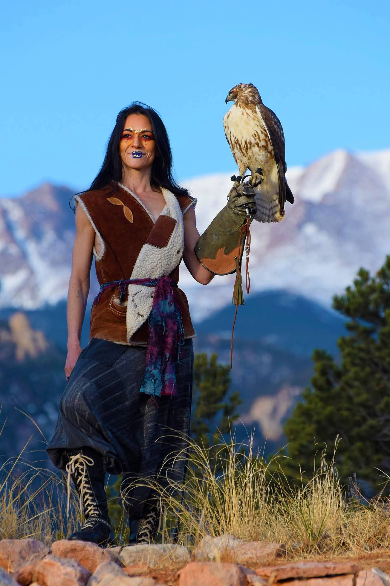 Person holding a bird of prey outdoors with mountains and trees in background, wearing traditional clothing.