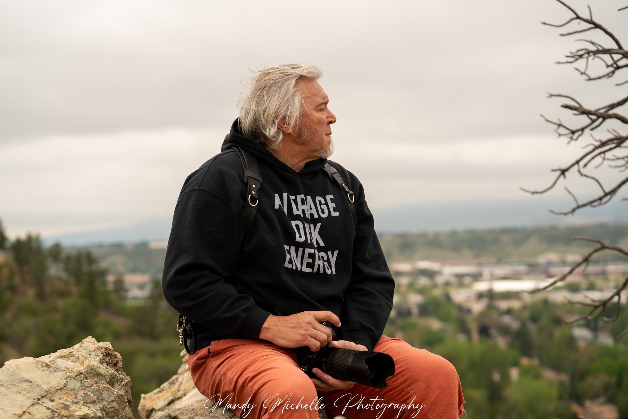 Older man with white hair sitting outdoors on a rock, wearing a black jacket and orange pants, with a landscape background.