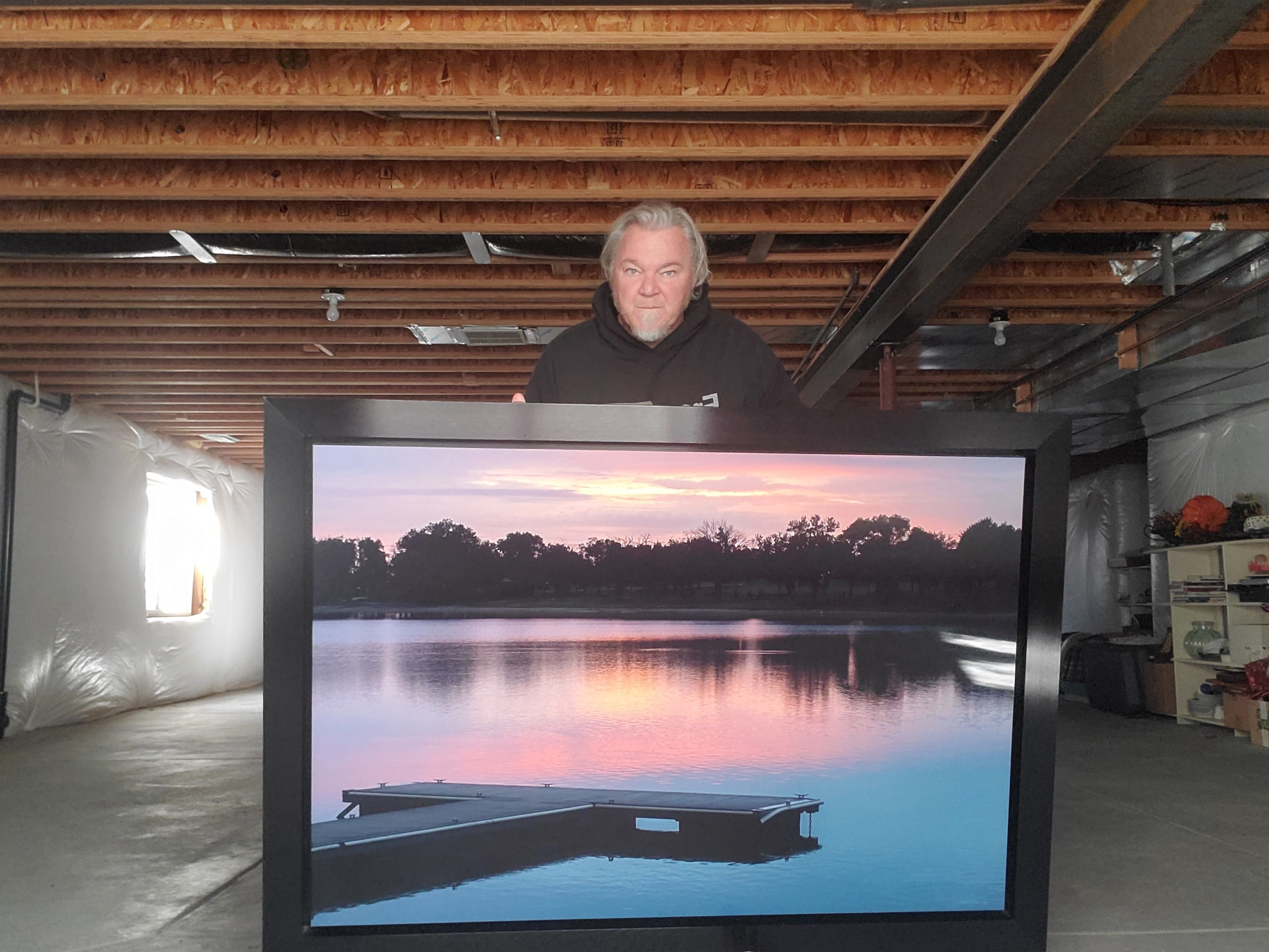 Person standing behind a large framed photograph of a lake with a boat, trees, and a sunset sky, in a basement setting.