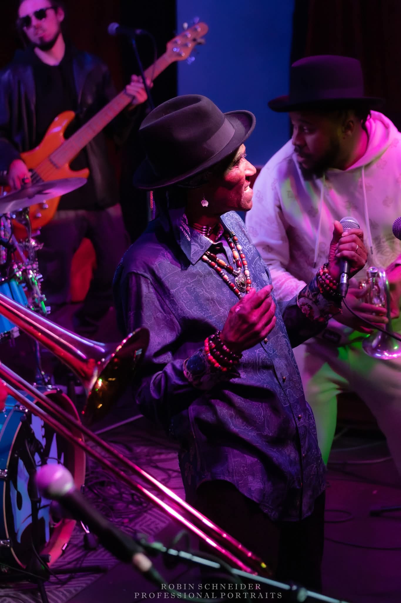 Two women in hats and jewelry talking on stage with musicians and instruments in background.