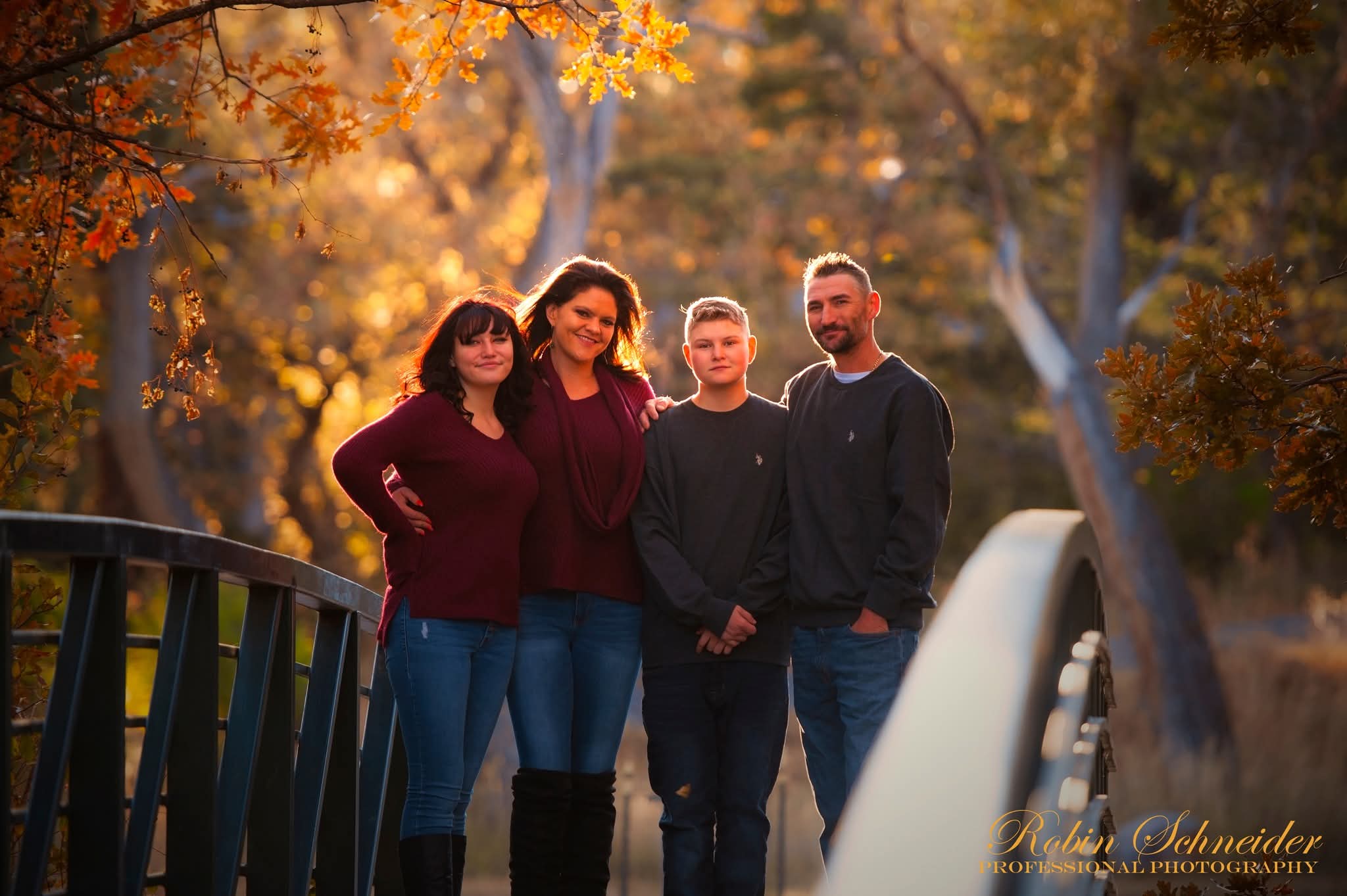 Group of four people standing on a bridge outdoors during autumn, trees with fall foliage in background.