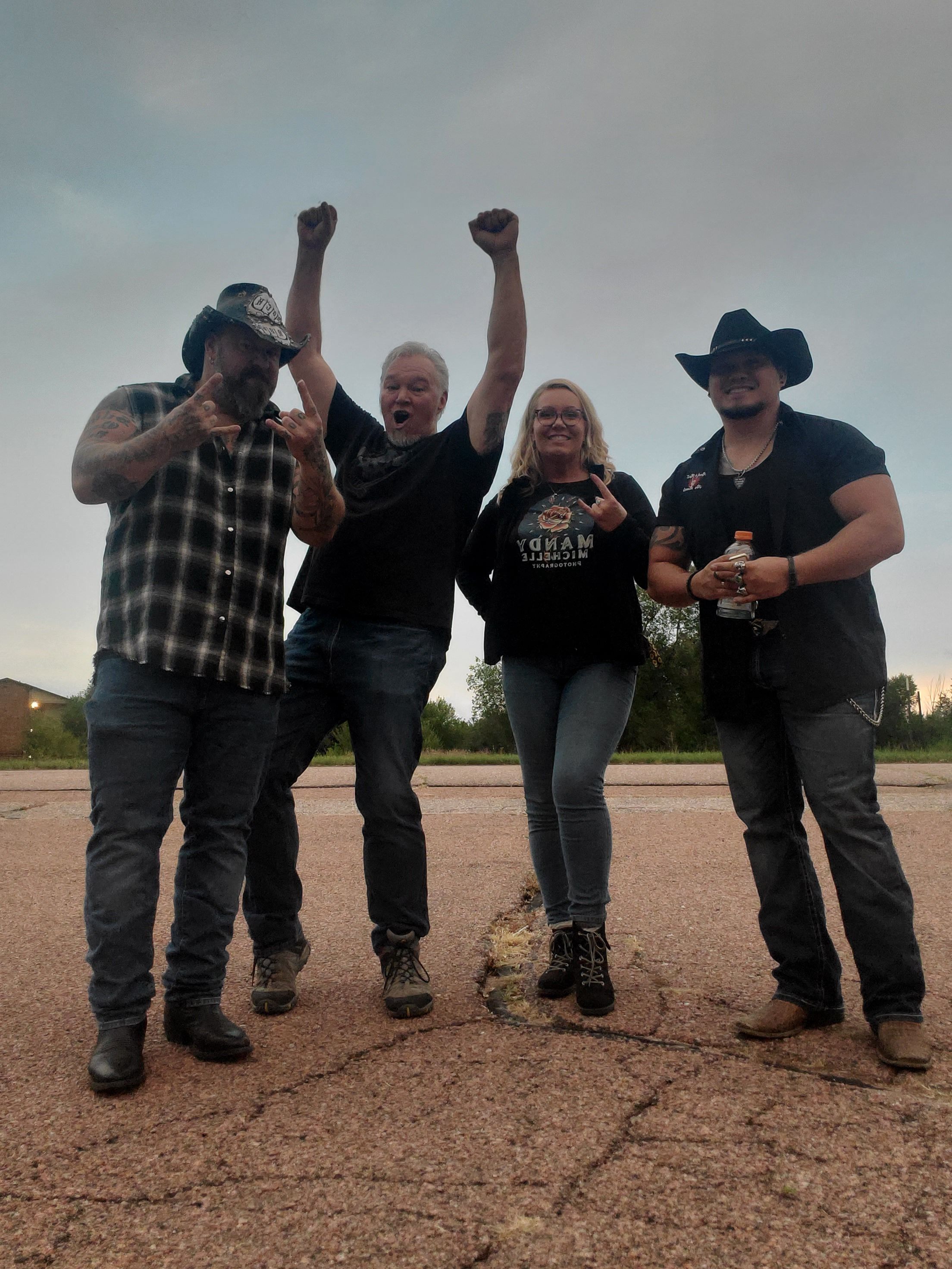 Four people standing outdoors on a dirt path, some raising fists, wearing casual clothing and hats.