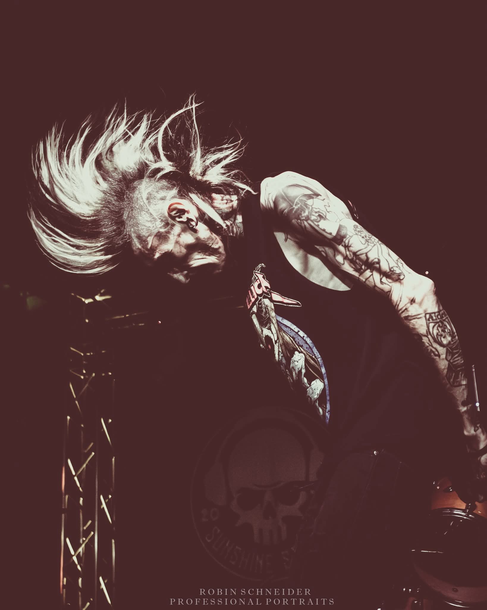 Person with long, flowing hair and tattoos, head tilted back, dark background, skull graphic on shirt, intense expression.