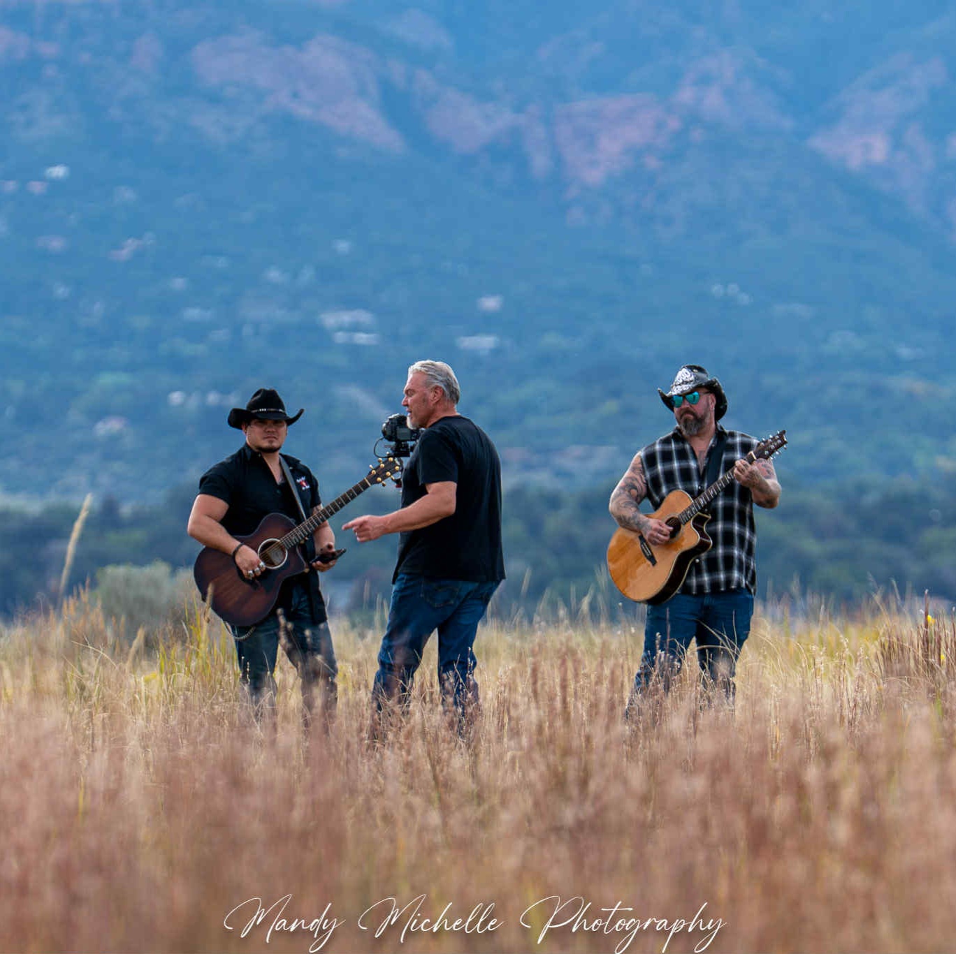 Three people playing guitars outdoors in a grassy field with mountains in the background.