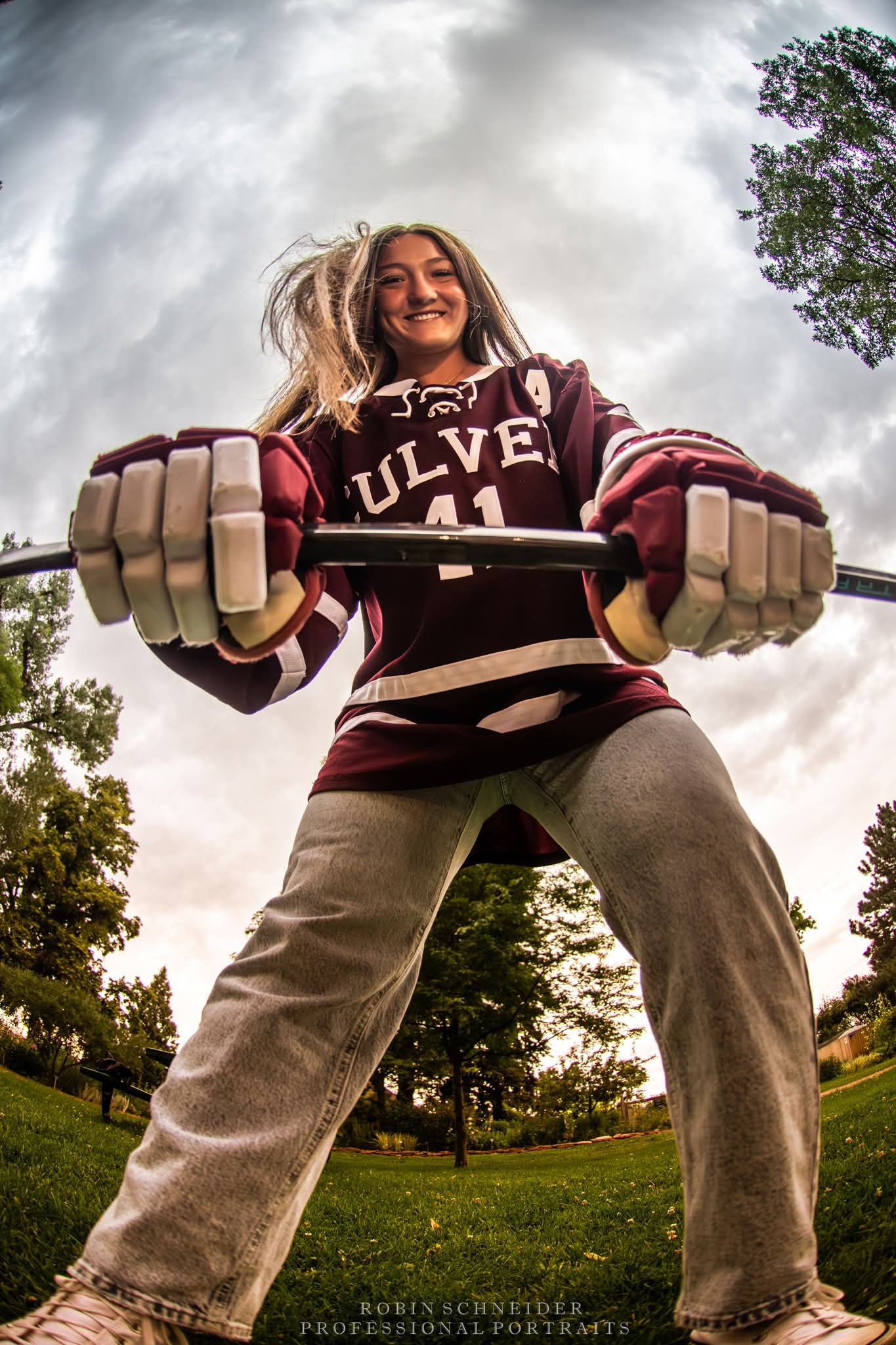 Person outdoors holding a hockey stick, smiling, wearing a maroon and white sports jersey, with trees and sky in background.