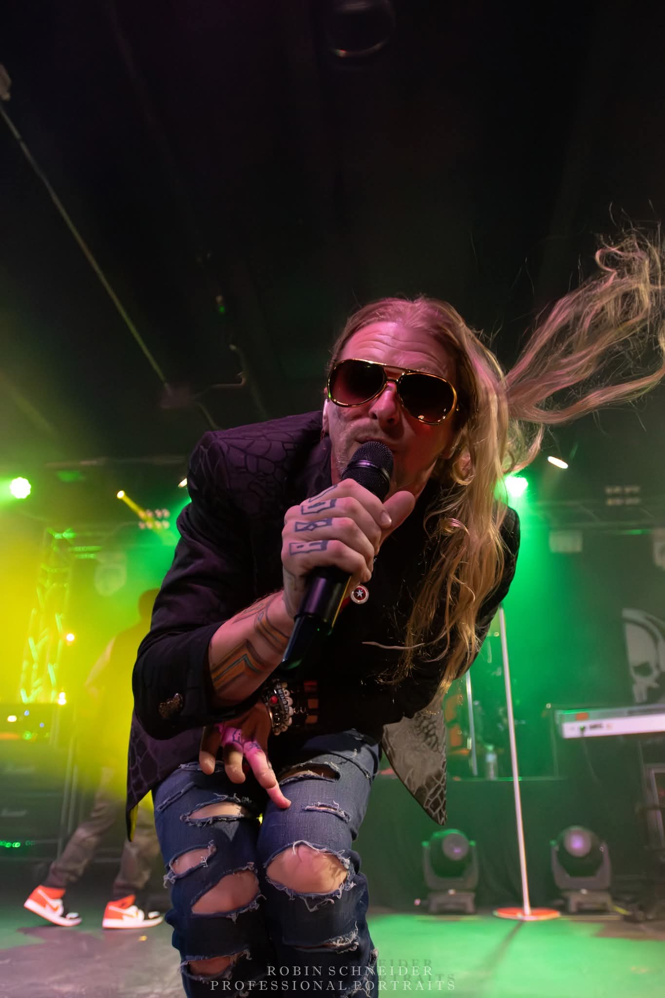 Female singer with long hair, sunglasses, and ripped jeans performs on stage with colorful lights.