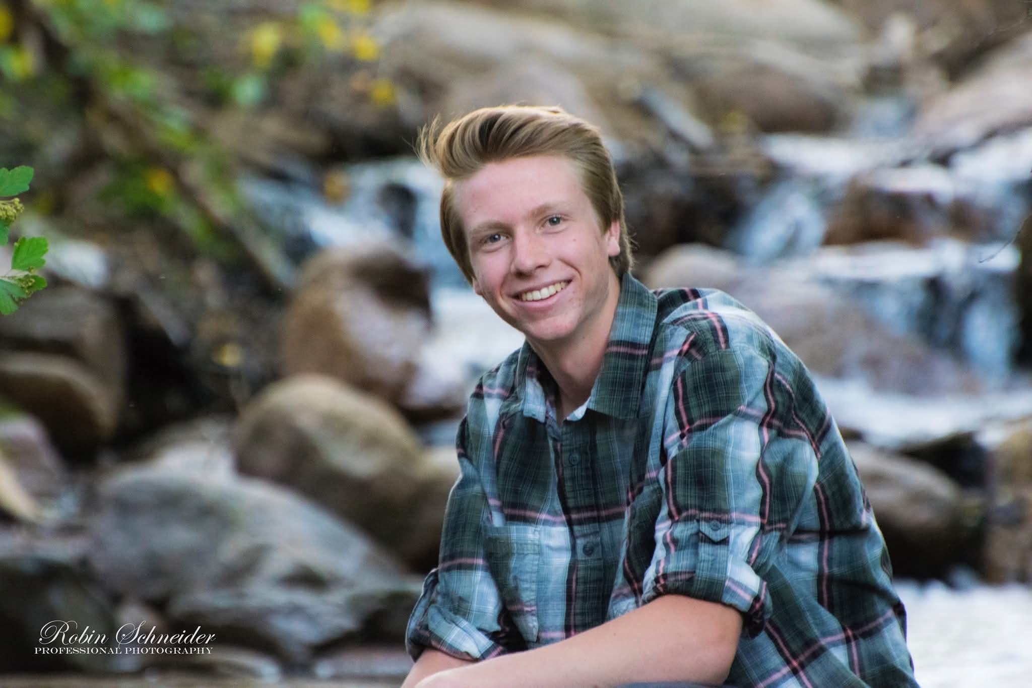 Young person smiling outdoors near a stream, wearing a plaid shirt, with rocks and water in the background.