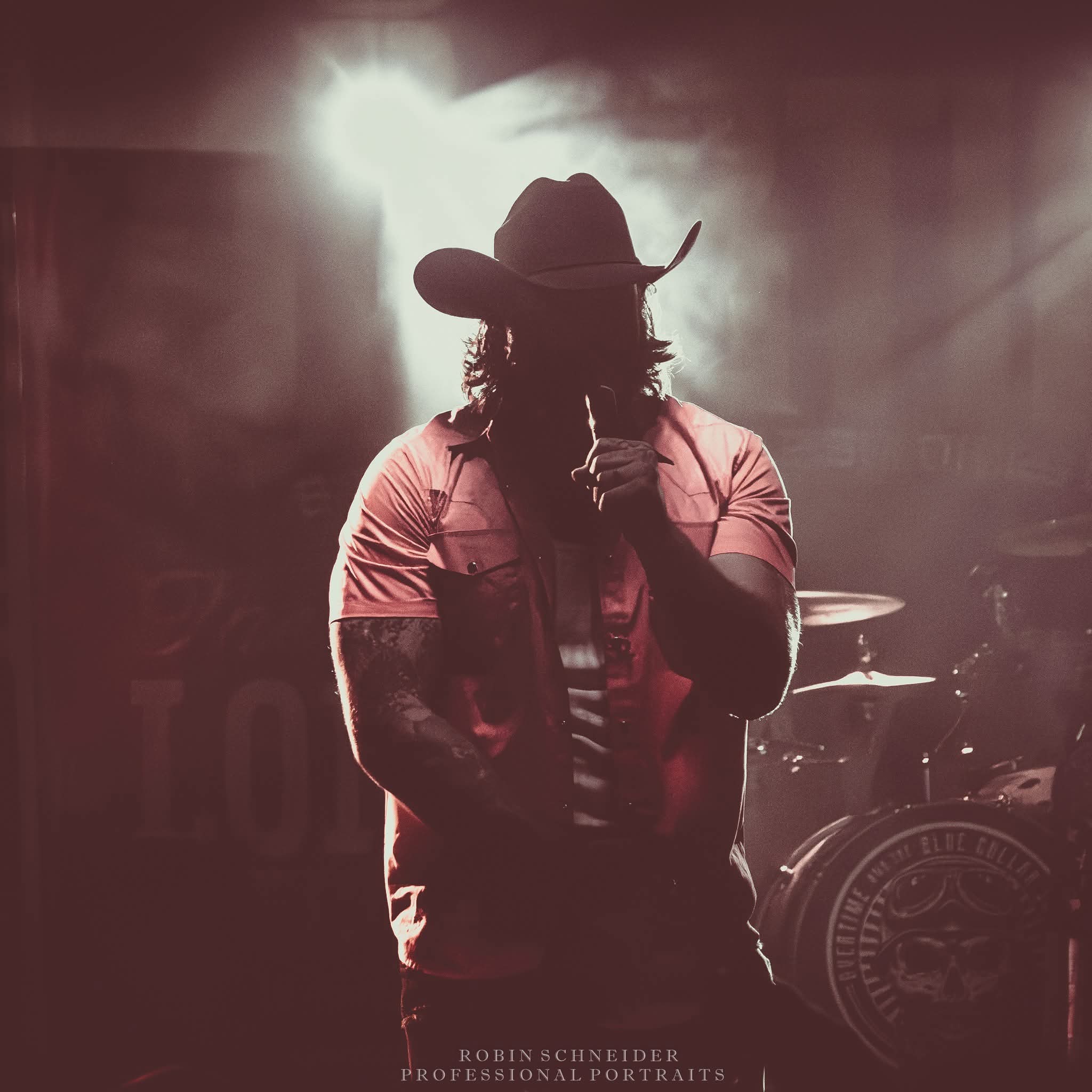 Person wearing a hat and striped shirt holding a microphone on stage with bright backlight, drums visible in background.