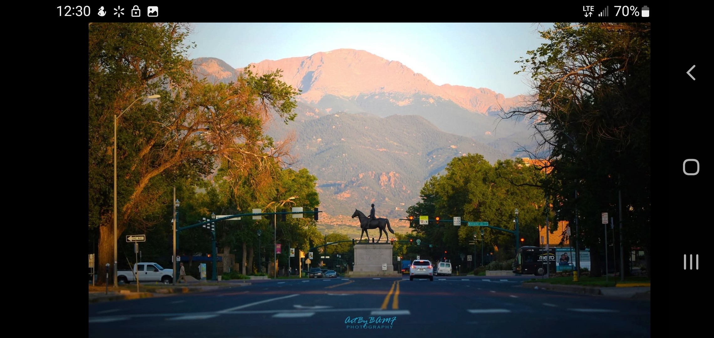 Street view with trees, cars, and a statue of a horse and rider, mountains in the background, and a clear sky.