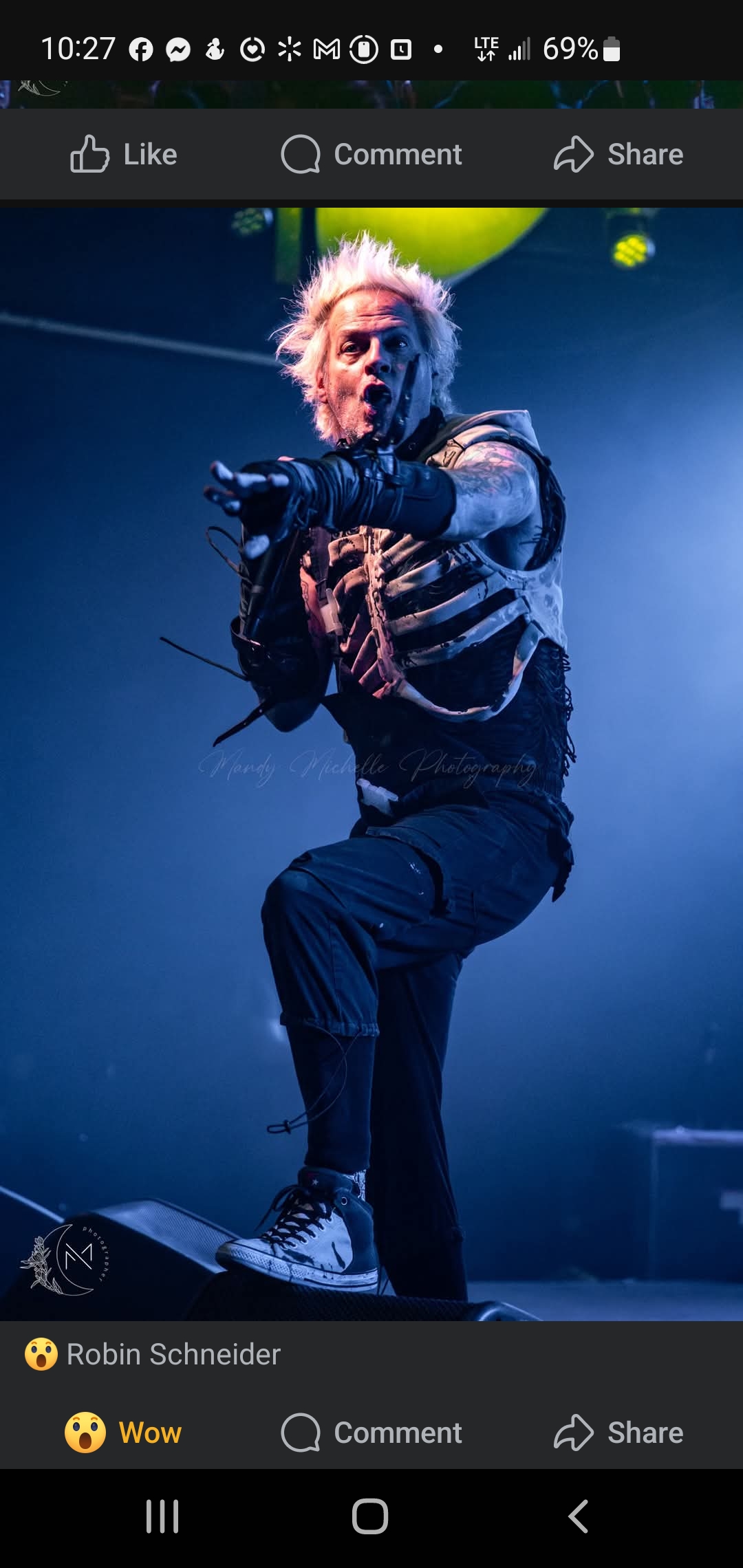 Performer with spiky hair and striped shirt on stage, holding a microphone, with blue lighting in the background.