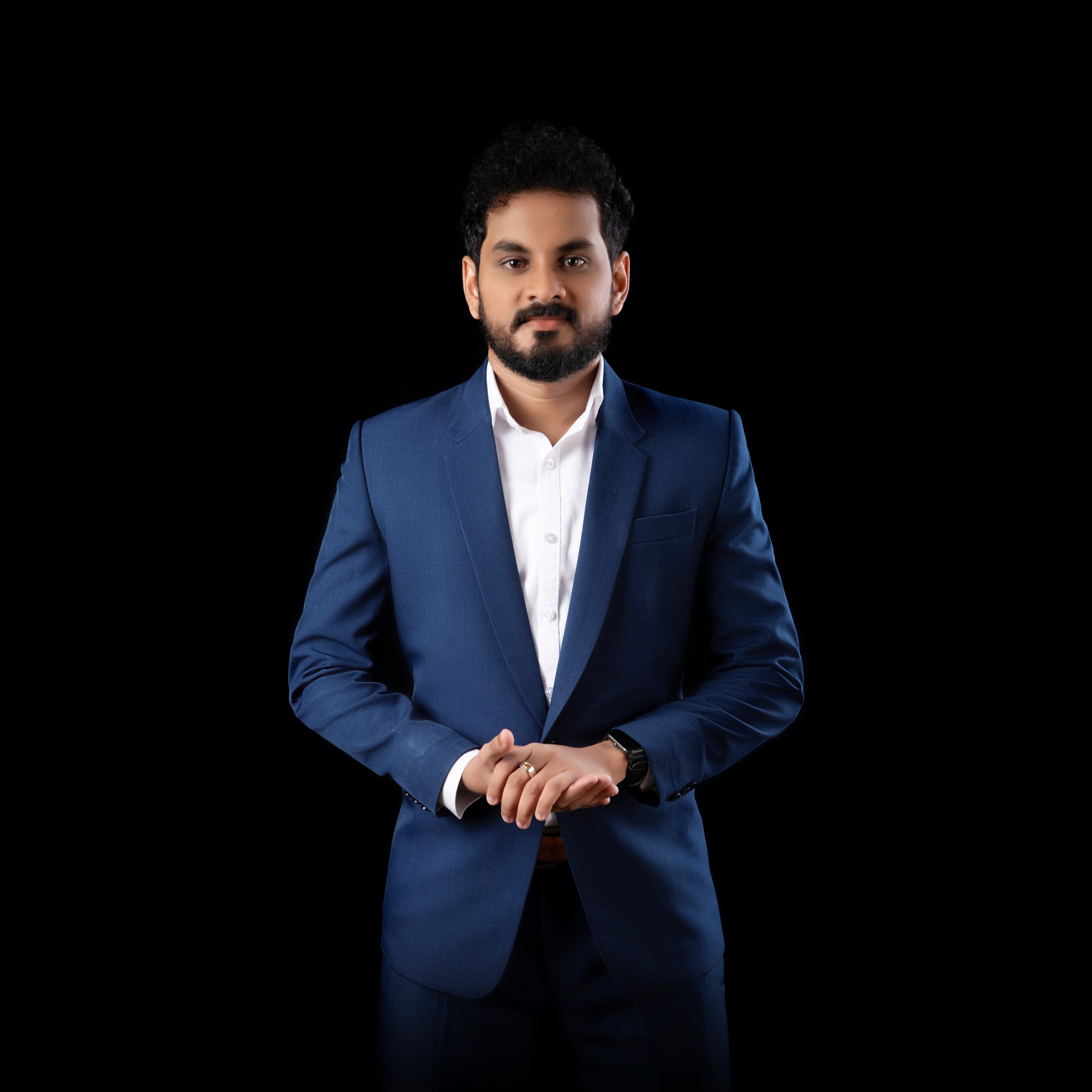 Man in a blue suit and white shirt standing with hands clasped against a black background.