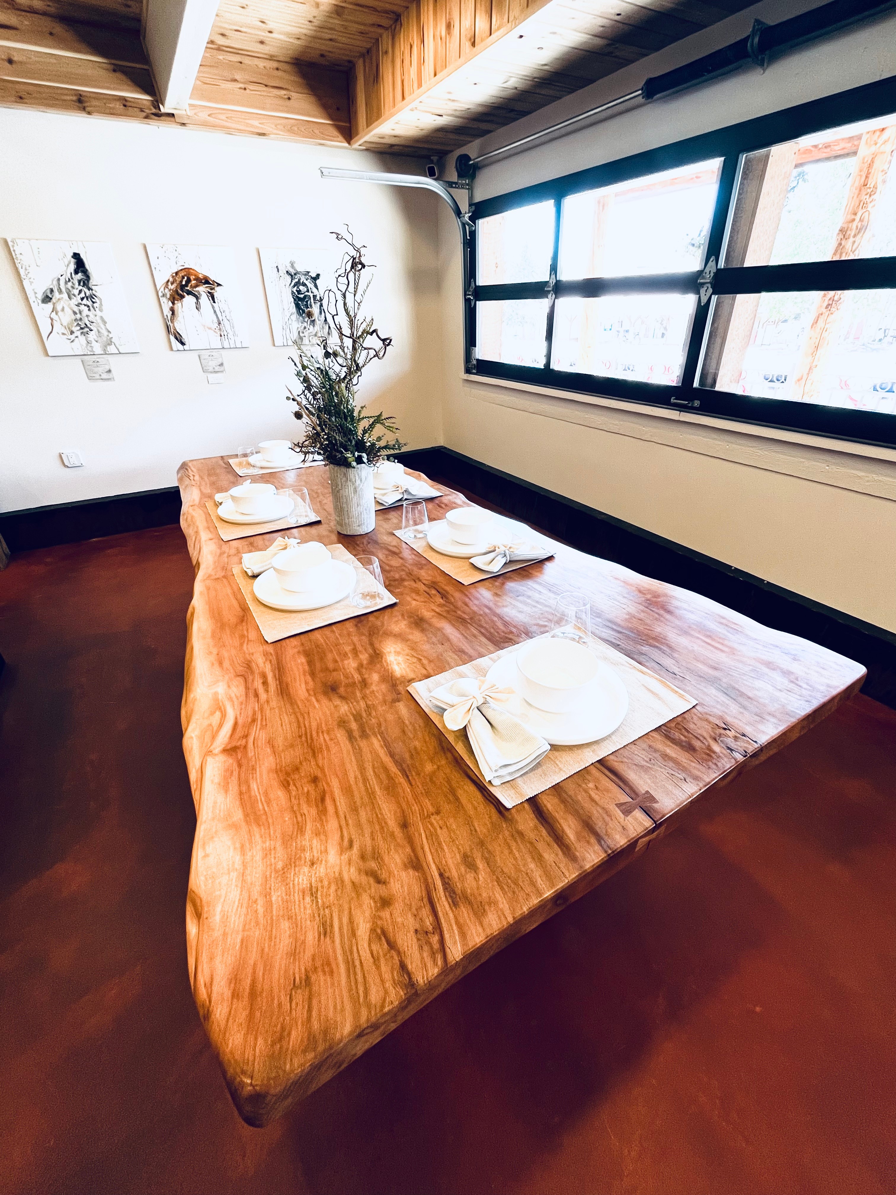 Wooden dining table set with plates, napkins, and a floral centerpiece, near windows with natural light.