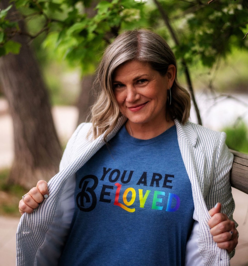 Woman outdoors wearing a blue shirt with rainbow-colored text, smiling, holding open a light-colored blazer, trees in background.
