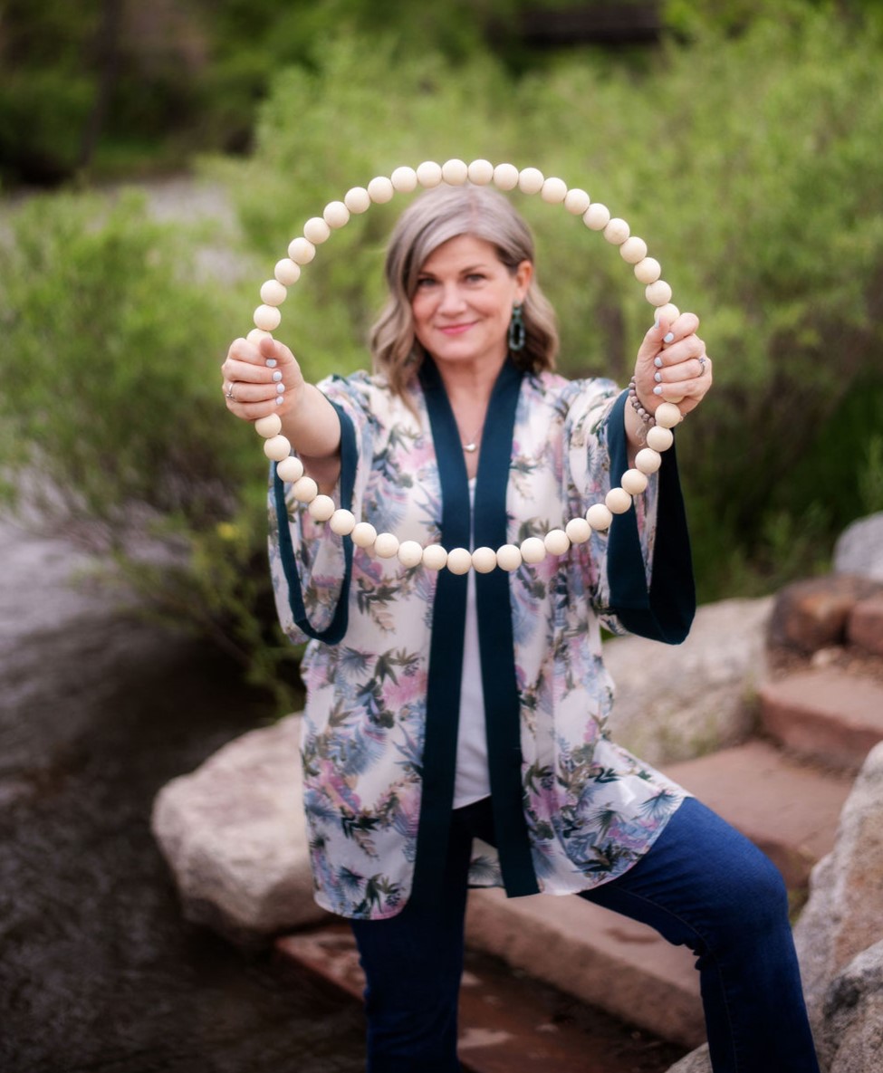 Woman outdoors holding a large circular bead necklace, smiling, with greenery and rocks in background.