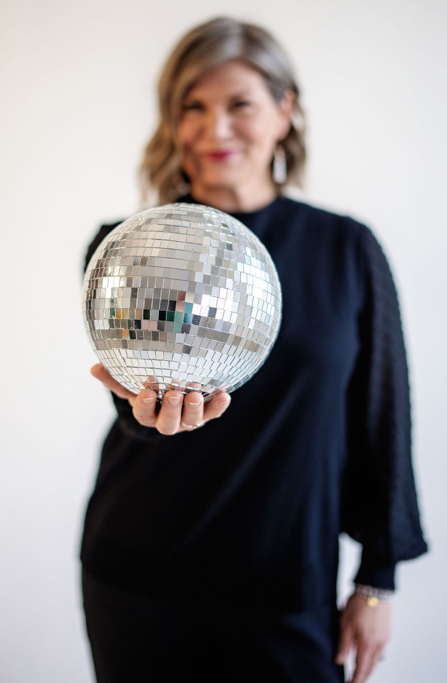 Woman holding a disco ball, smiling, with a blurred background, wearing a dark outfit.