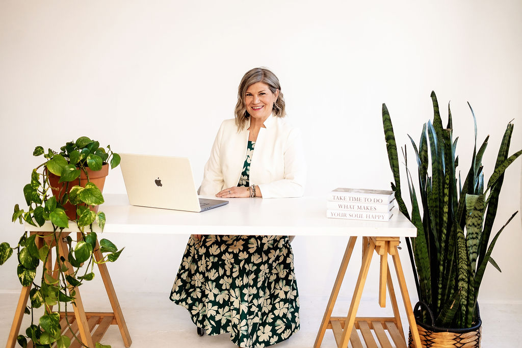 Woman sitting at a white desk with a laptop, surrounded by potted plants, smiling, in a bright room.