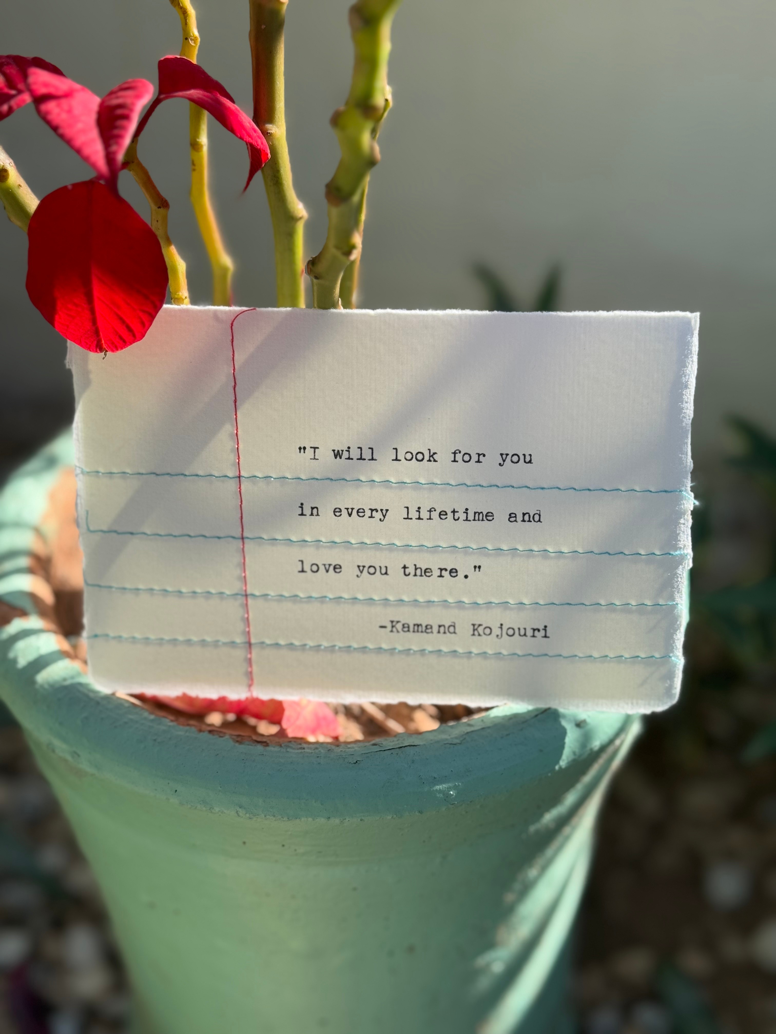 A white card with a quote, placed in a plant pot with pink flowers and green stems, outdoors.