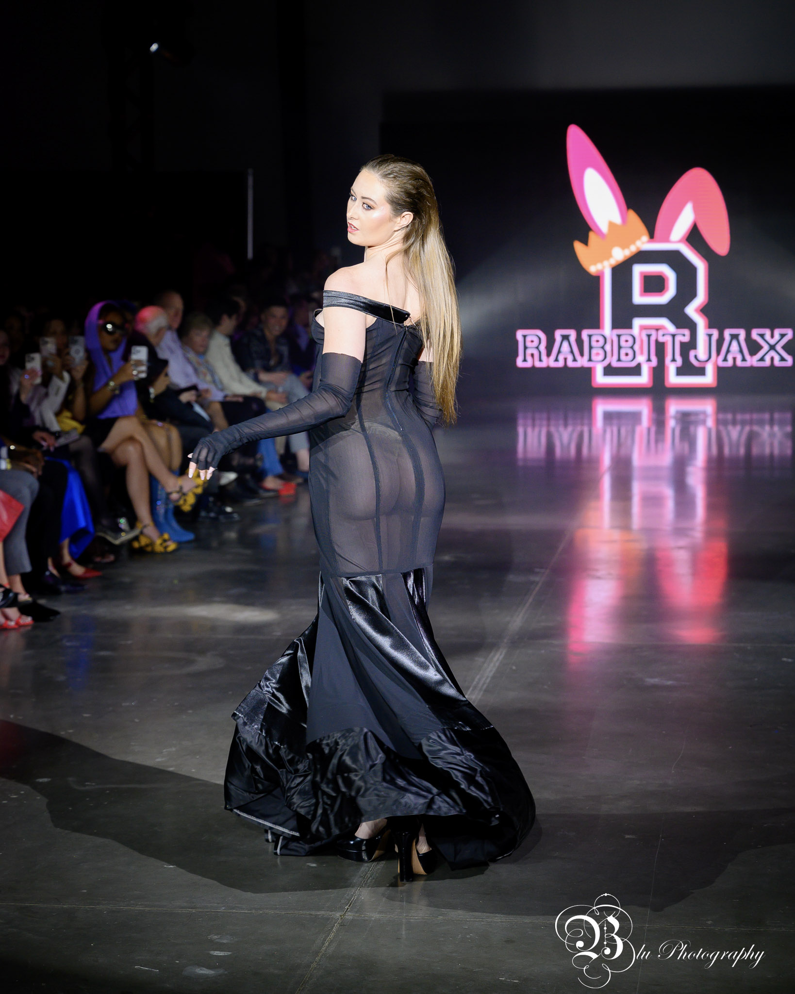 Model walking on runway in sheer black dress with long gloves, audience seated on sides, logo in background, dark setting.
