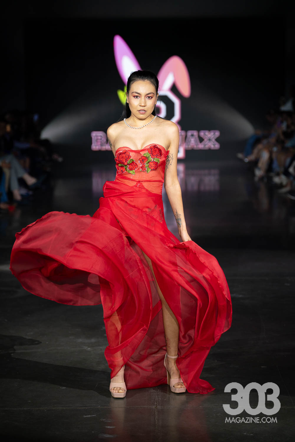 Woman in red dress walking on runway with a butterfly logo background, dark setting, audience on sides, confident pose