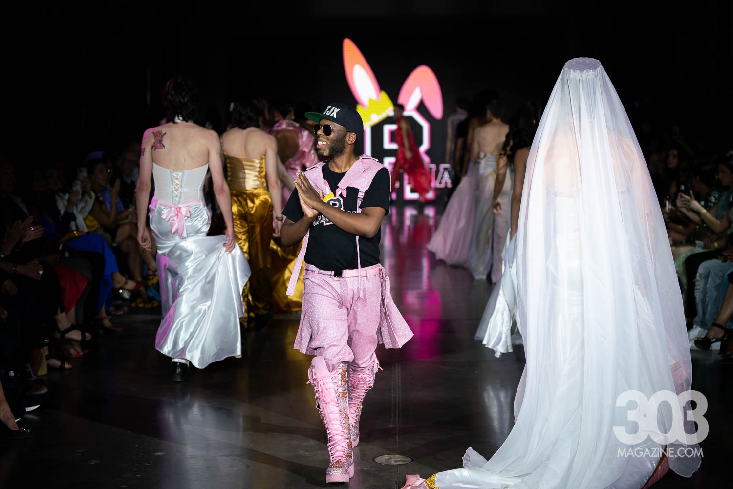 Person walking on runway with bunny ears headband, surrounded by models in dresses, audience seated, stage background with logo, dark lighting