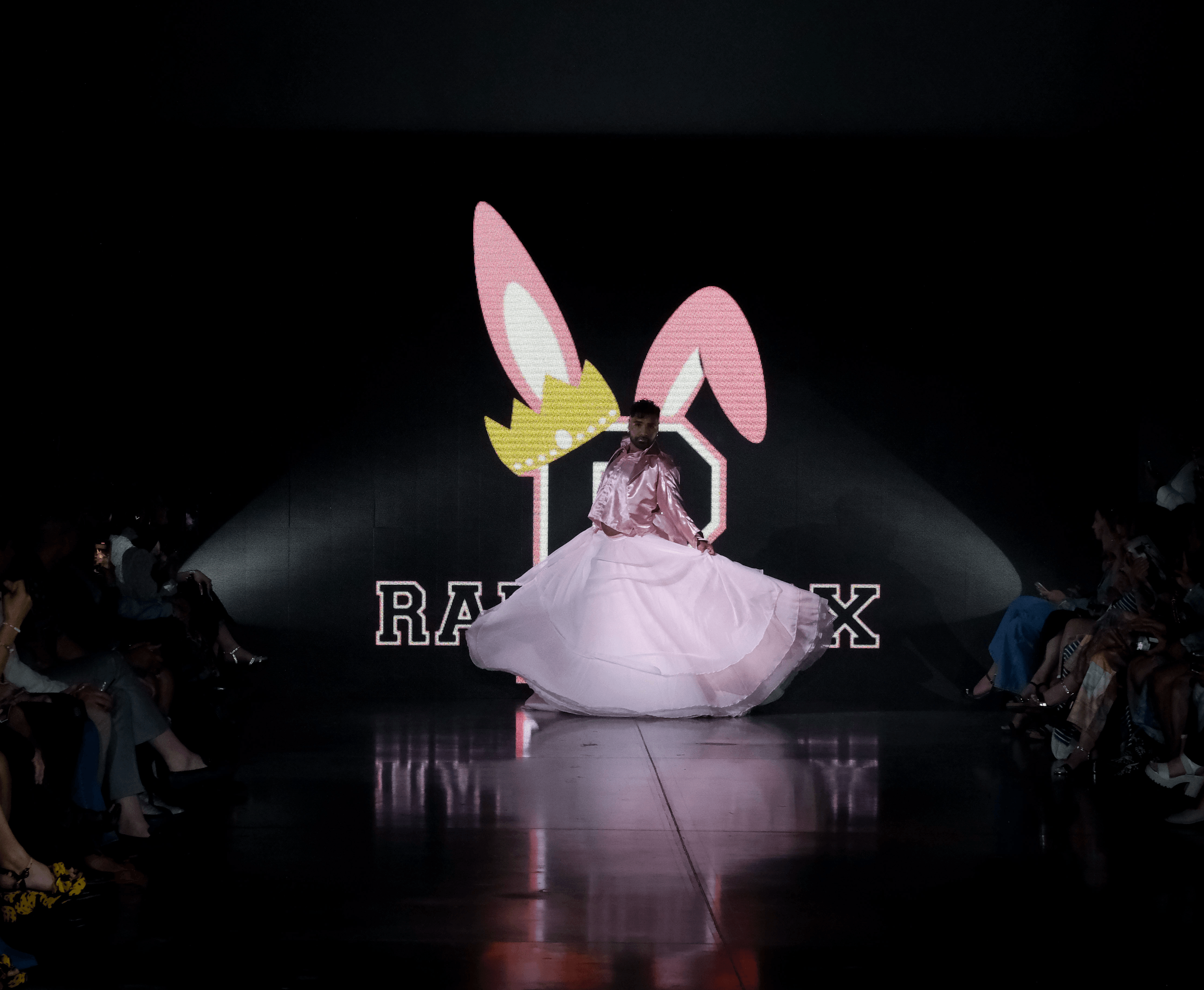 Person in pink dress with bunny ears on stage, large bunny ears graphic in background, audience seated on sides, dark setting.