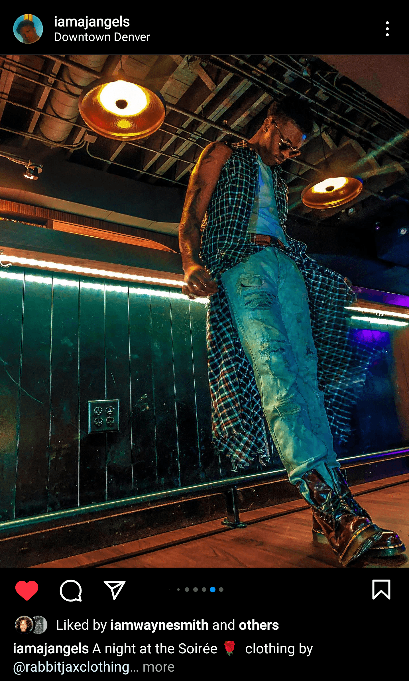 Person standing in a nightclub with colorful lighting, wearing a patterned outfit and jeans, leaning against a bar or railing.