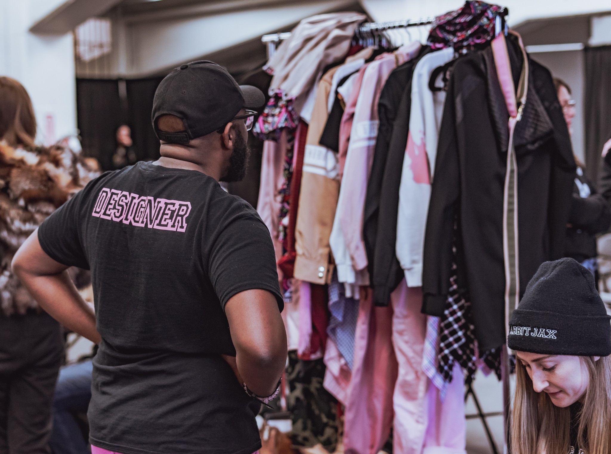 Person wearing a black shirt with 'DESIGNER' on the back shopping for clothes at a market stall.