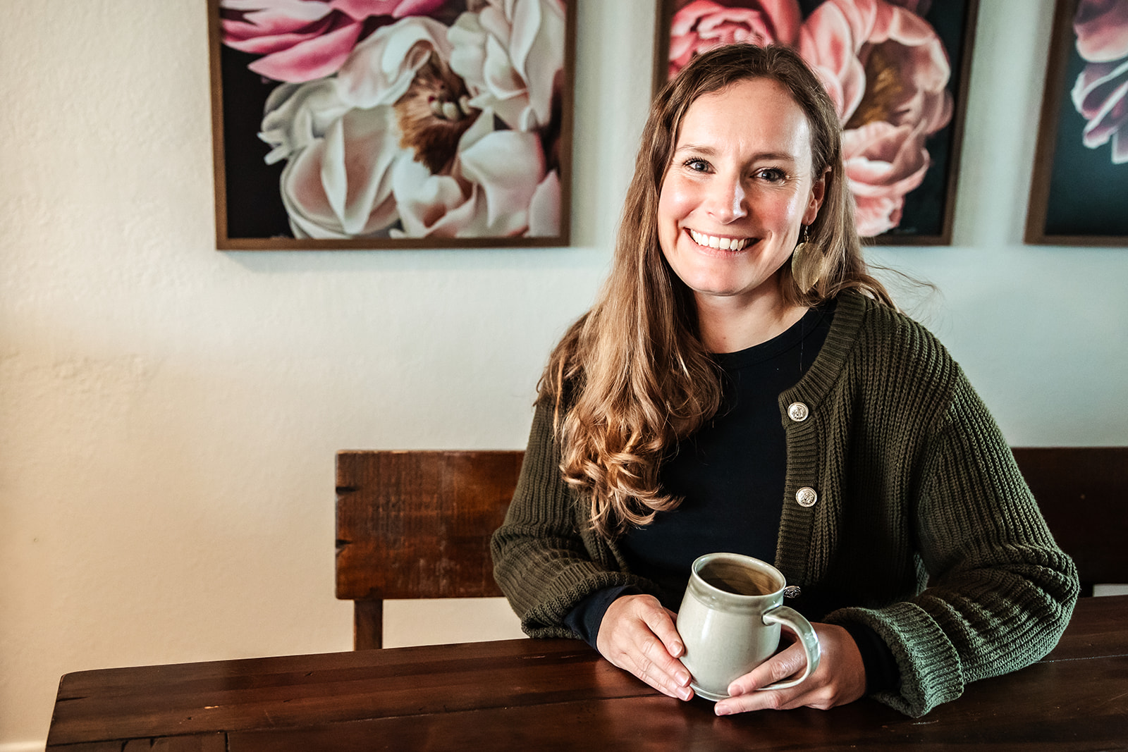 Smiling woman with long hair sitting at a table holding a mug, with floral artwork on the wall behind her.