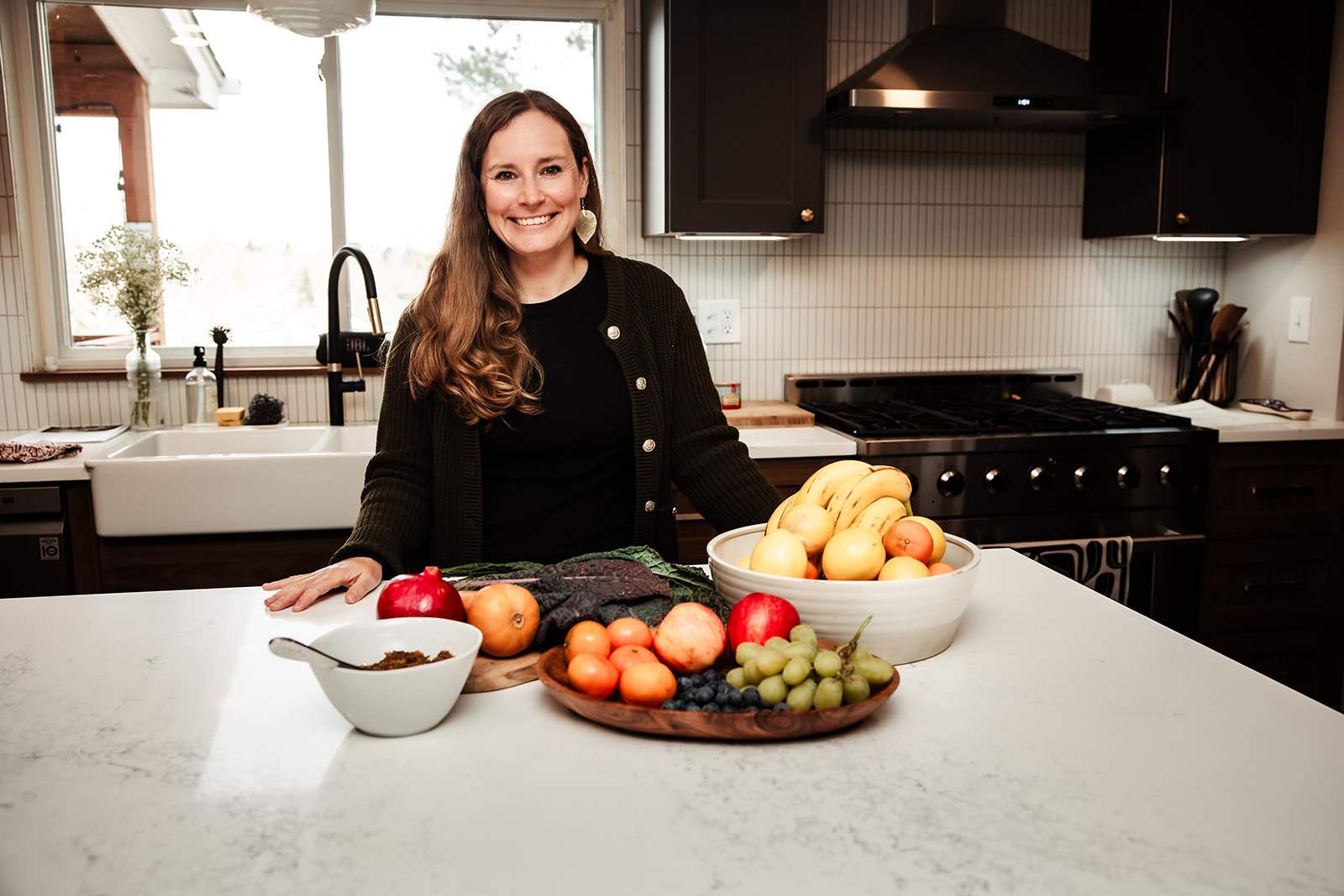 Woman standing in kitchen with bowls of apples, peaches, grapes, and vegetables on white countertop.