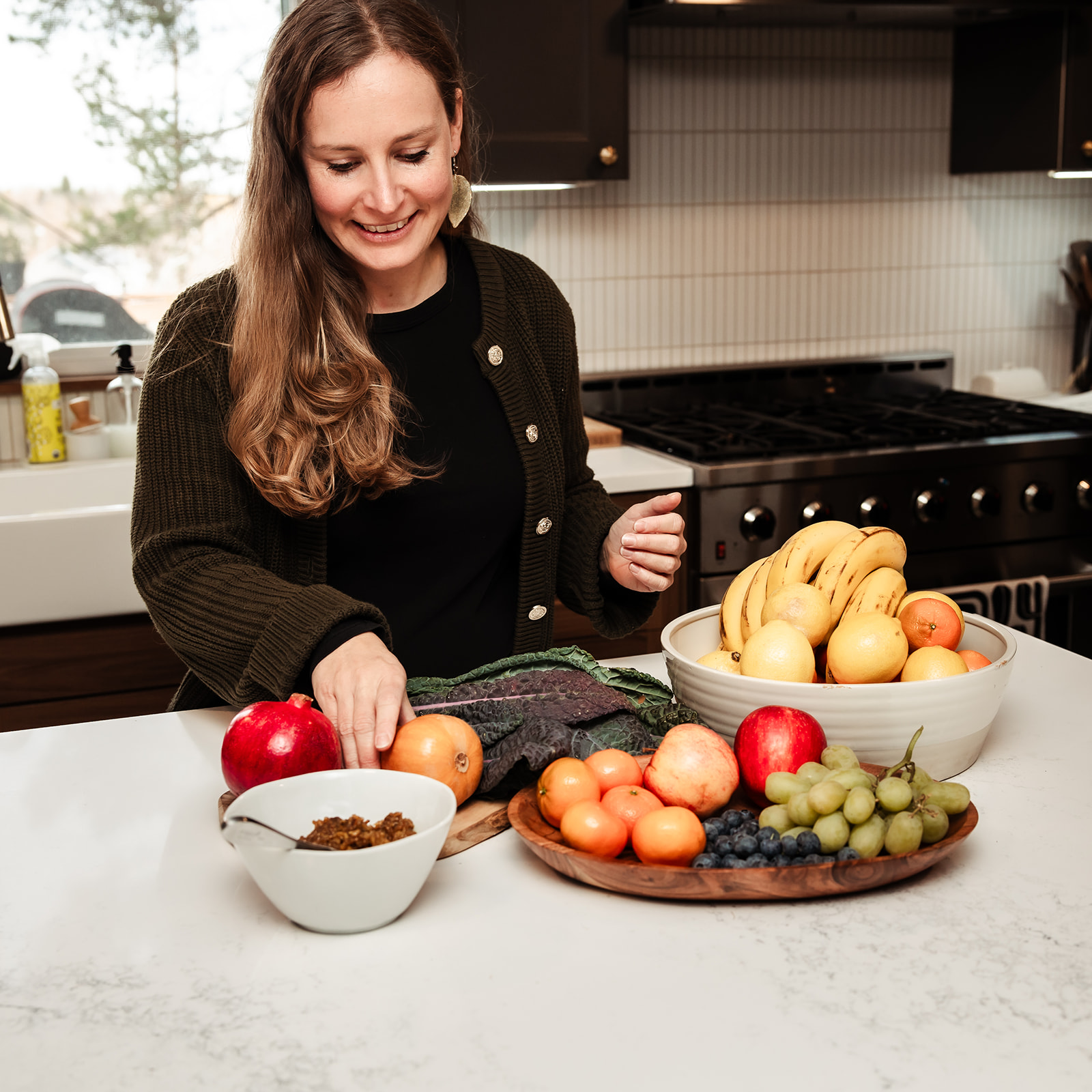 Woman smiling at a kitchen counter with bowls of apples, grapes, and bananas, and a plate of peaches and tomatoes.