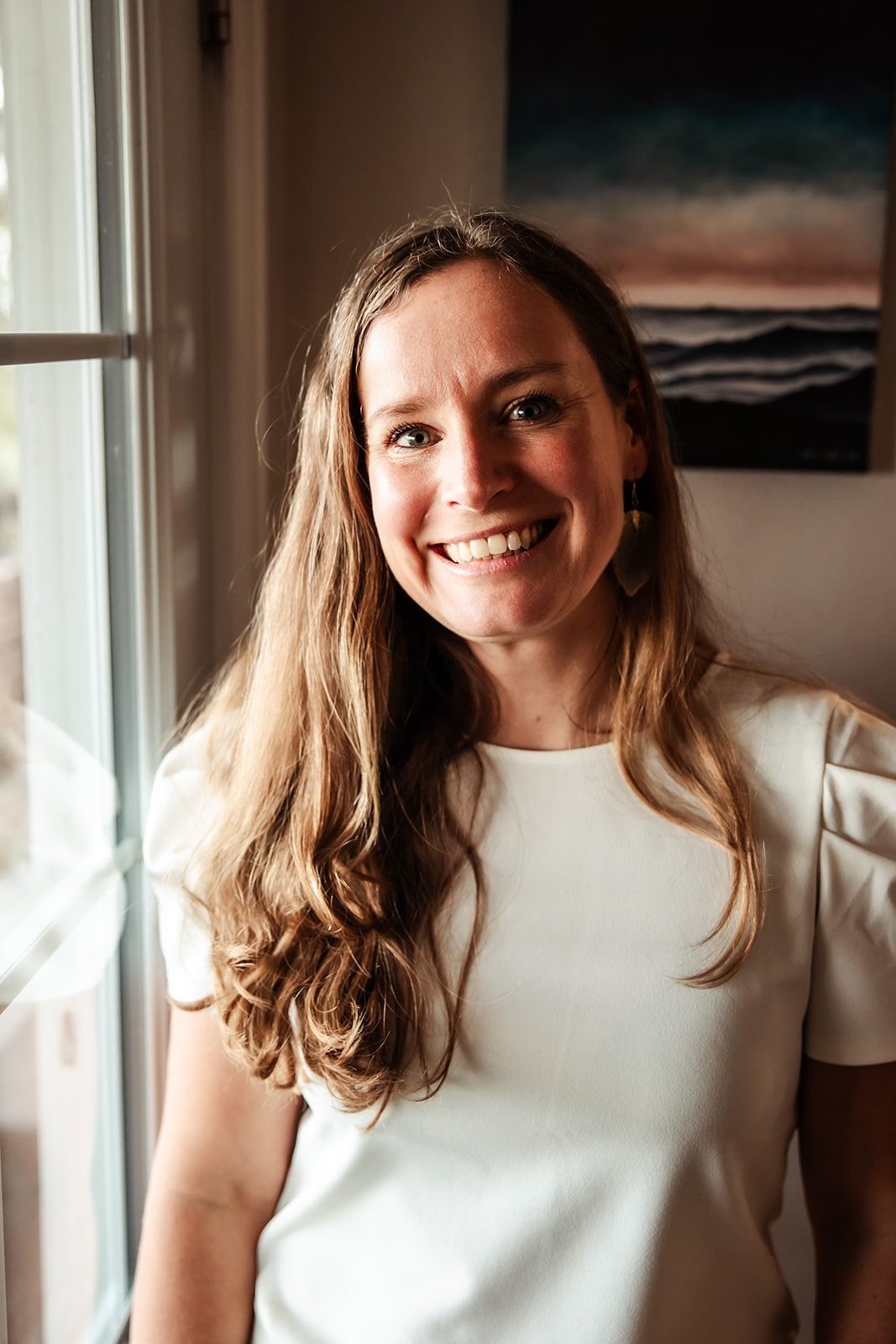 Smiling woman with long wavy hair standing near a window, wearing a white top, with a painting in the background.