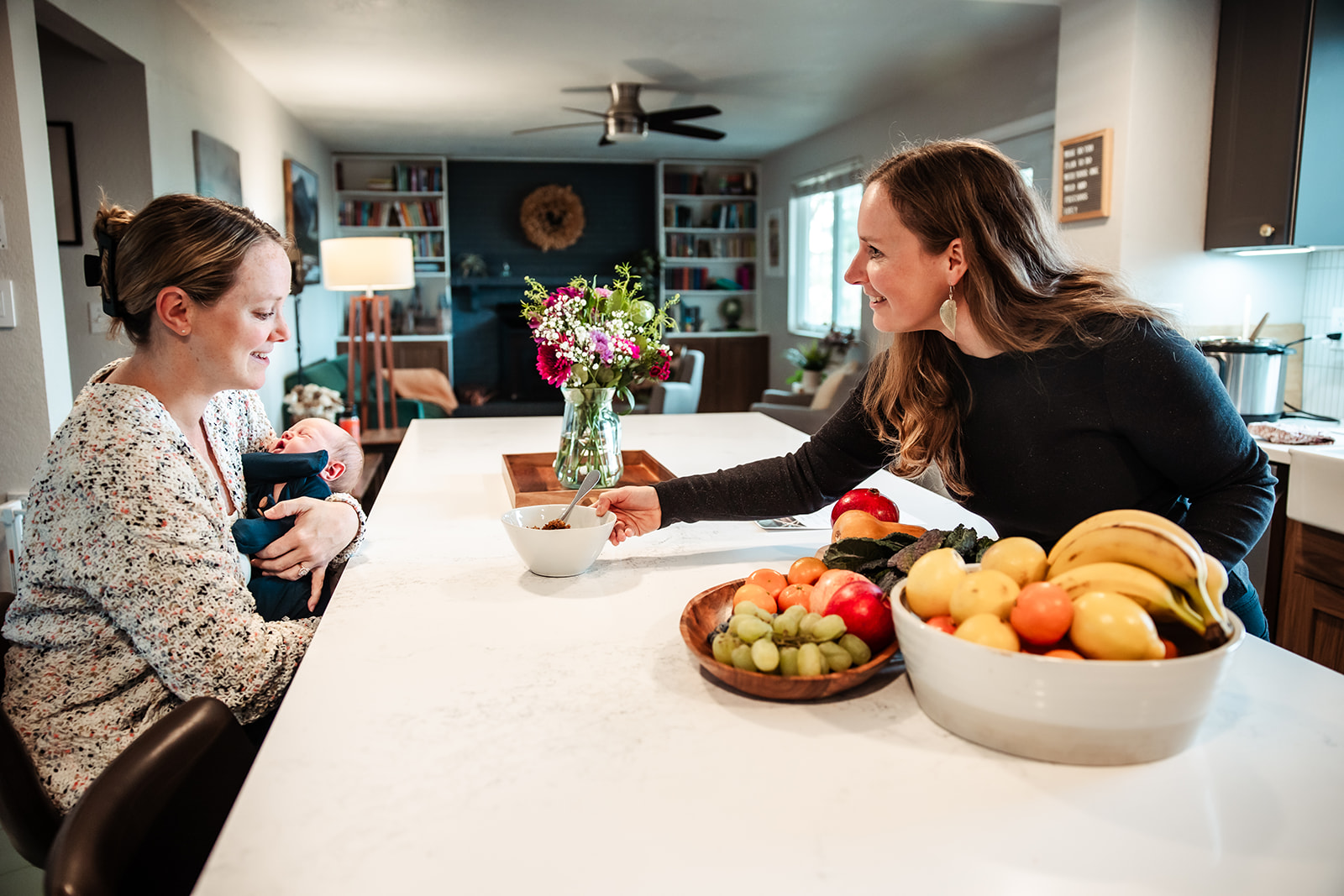 Two women exchange gifts at a kitchen island with bowls of fruit and a vase of flowers, smiling at each other.