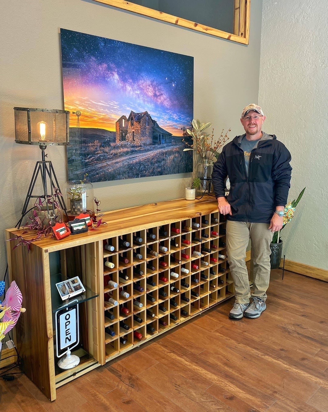 Person standing next to a wooden wine rack with a large screen displaying a landscape behind them.