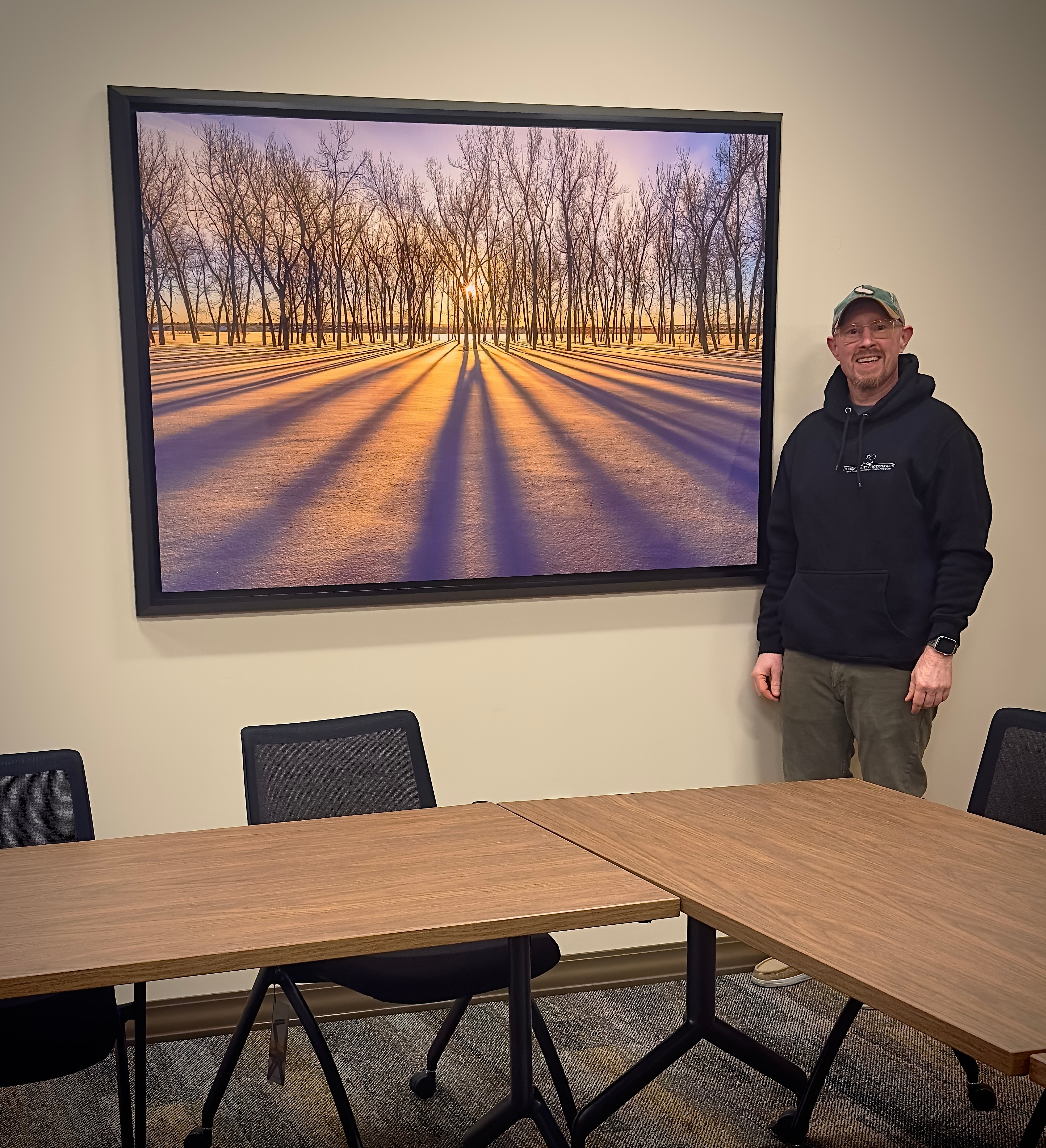 Man standing next to a large framed landscape photograph of trees and shadows on snow, in a conference room with a table and chairs.