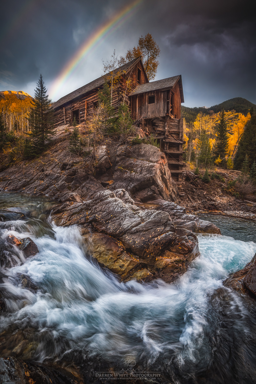 A rustic wooden cabin on a rocky riverbank with a rainbow in a cloudy sky and autumn trees in the background.