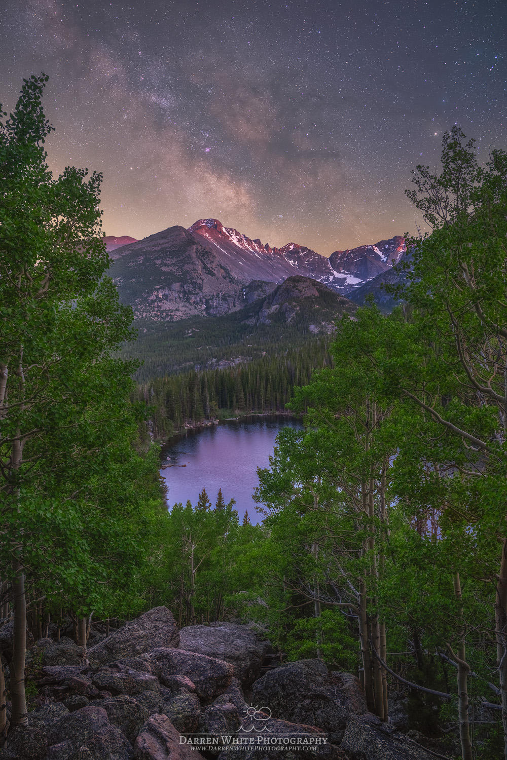 Night sky with stars above mountain range and a lake surrounded by trees in foreground.