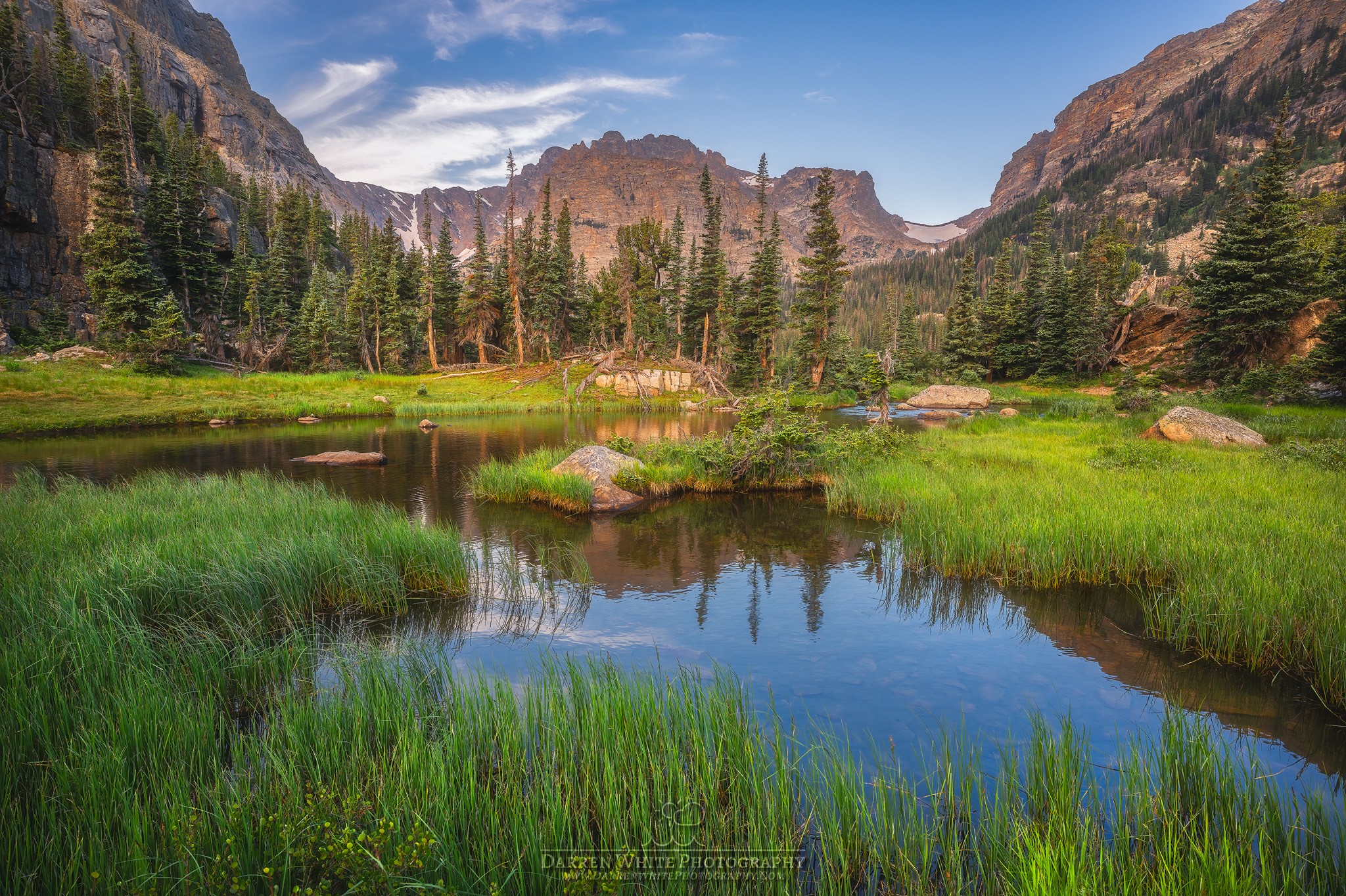 Mountain landscape with a lake, green grass, trees, and rocky mountains in the background.