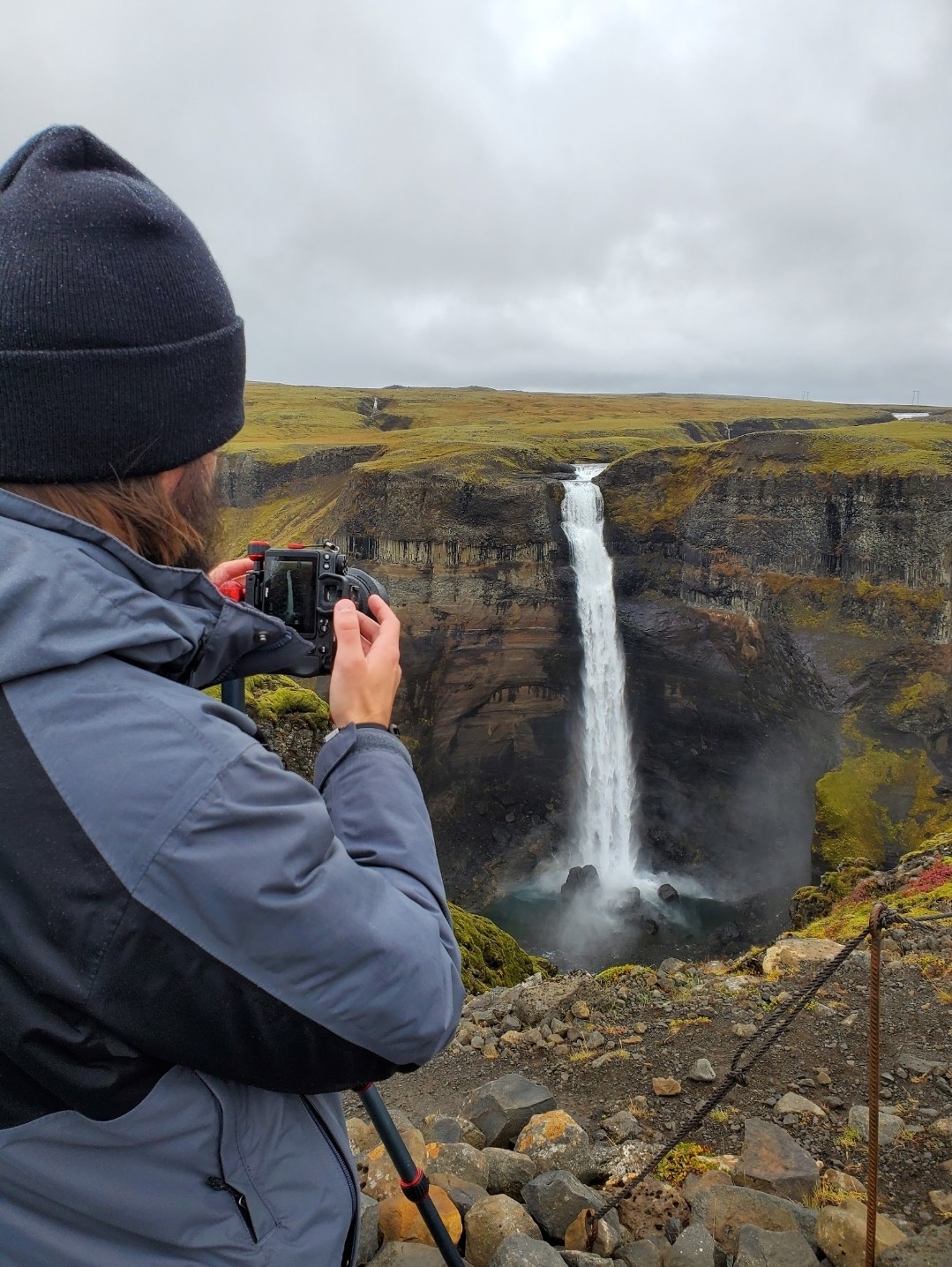 Person in a dark beanie and gray jacket taking a photo of a waterfall on a cloudy day, with rocky ground in foreground.