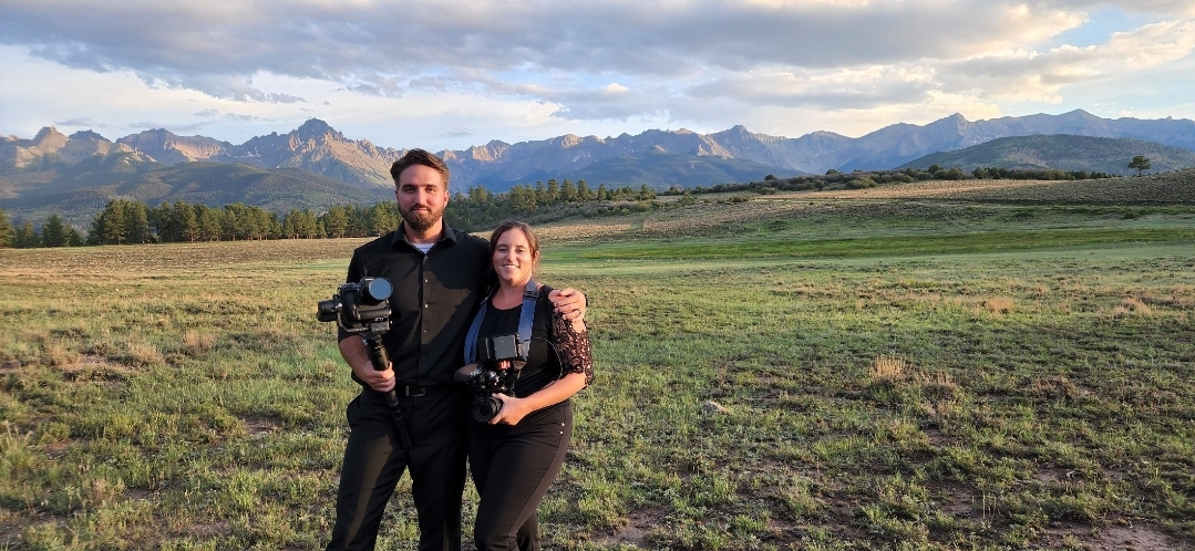 Two people standing outdoors with mountains and sky in background, holding cameras, smiling, in a grassy field.