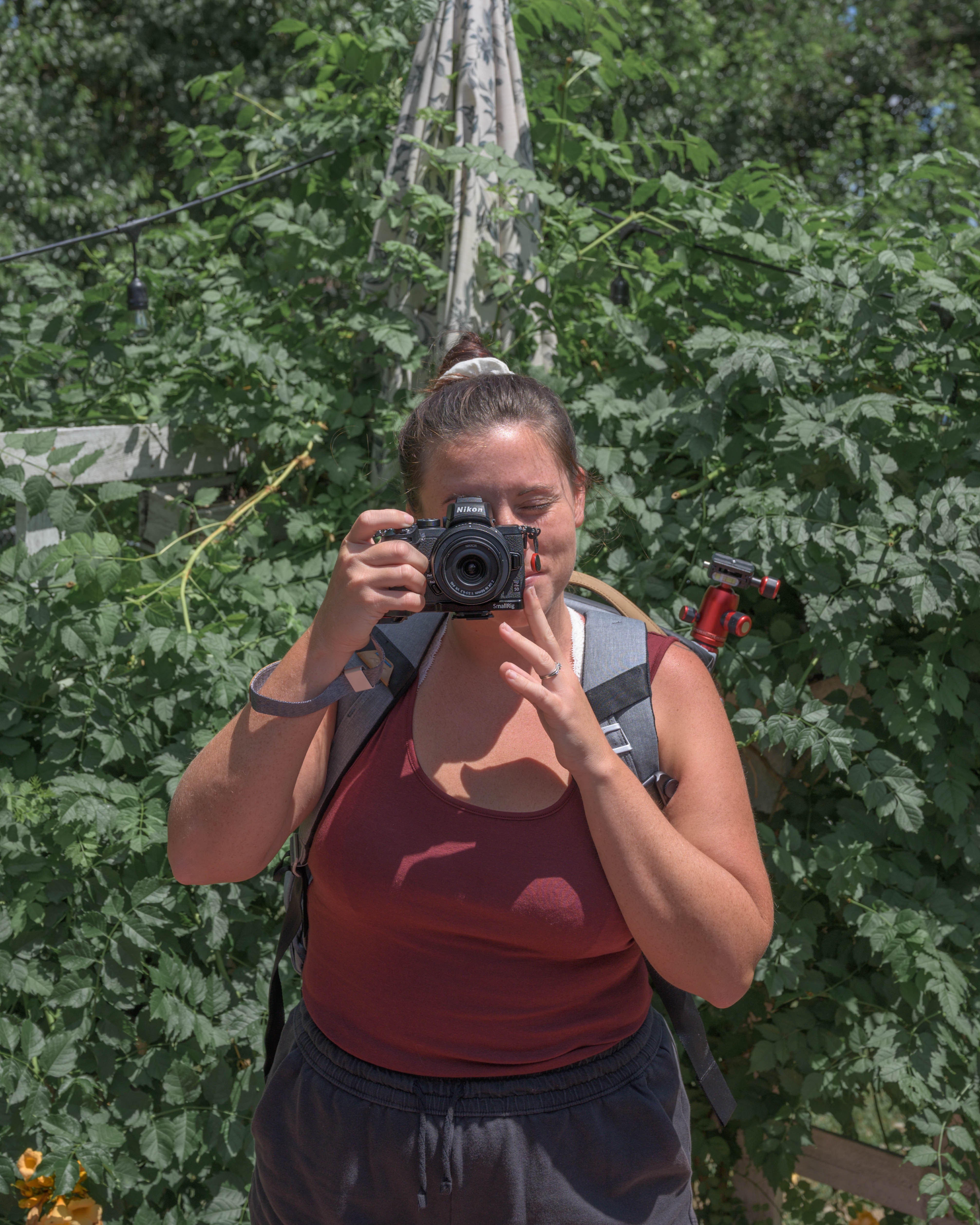 Woman taking a photo with a camera outdoors surrounded by green foliage, wearing a sleeveless top and glasses.
